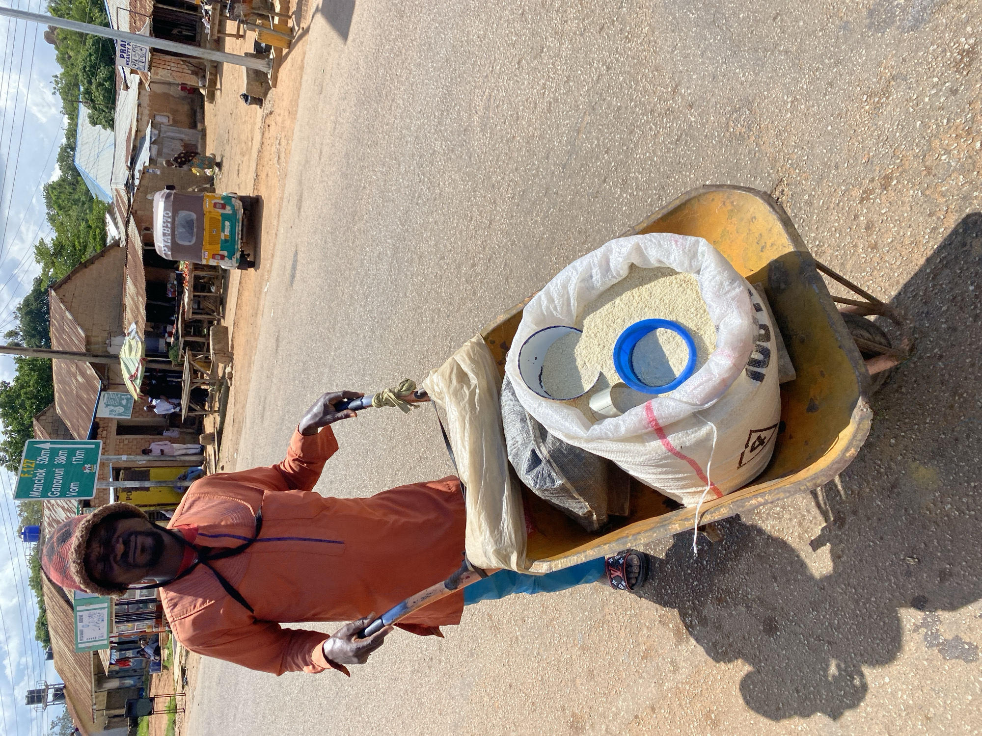 A man, wearing an orange tunic and a patterned hat, pushes a wheelbarrow filled with sacks of rice and other goods down a dusty street in Bukuru, Nigeria. The wheelbarrow also contains a white bag with red stripes, a dark sack, and a blue scoop. The man, with his eyes closed and a neutral expression, is the primary subject. He is wearing blue trousers and sandals. In the background, there are small shops with corrugated iron roofs, some with goods displayed outside. A green road sign indicates distances to Mancho, Ganawuri, and Vom. A yellow and green auto-rickshaw is parked on the right side of the street. The sky is partly cloudy. The scene depicts a commercial street in a Nigerian town.