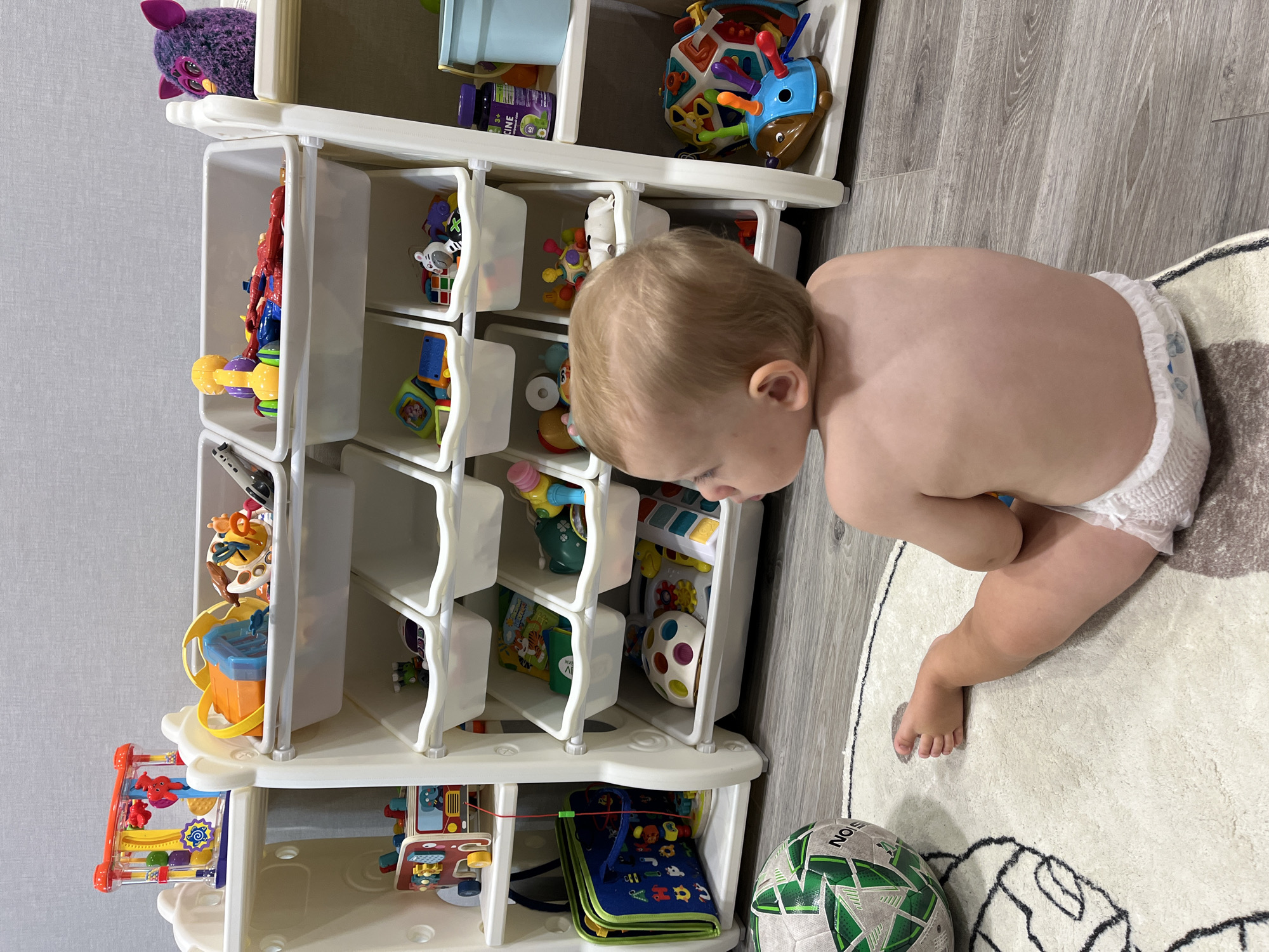 In a domestic setting, a diaper-clad toddler with blond hair sits on a circular rug featuring an abstract outline of a face. The toddler is facing away from the camera, looking towards a white plastic toy organizer filled with various toys. To the left of the toddler, a green and white soccer ball rests on the rug. The toy organizer has multiple tiers with individual bins. The bins contain an assortment of colorful children's toys, including blocks, stuffed animals, and developmental toys. The environment appears to be a child's play area, with a gray wall in the background and a wood-grain floor. This scene is not located in Nizhny Novgorod, Russia.