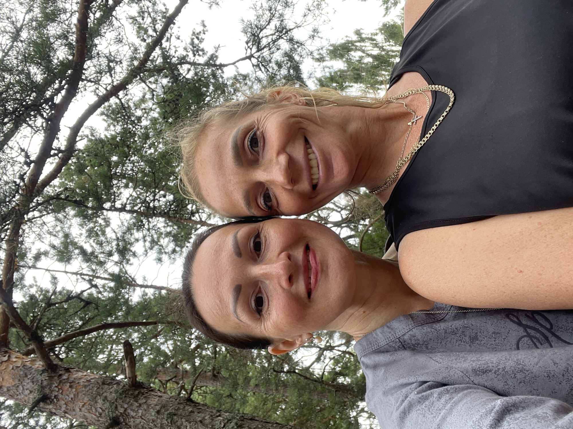 Two women are taking a selfie outdoors. The woman on the left has dark hair pulled back and is wearing a grey top. The woman on the right has blonde hair and is wearing a black tank top and gold necklaces. They are both smiling and their faces are close together. They are surrounded by trees, suggesting a park or forest setting. The image appears to be taken in Zelenaya Roshcha, Russia.