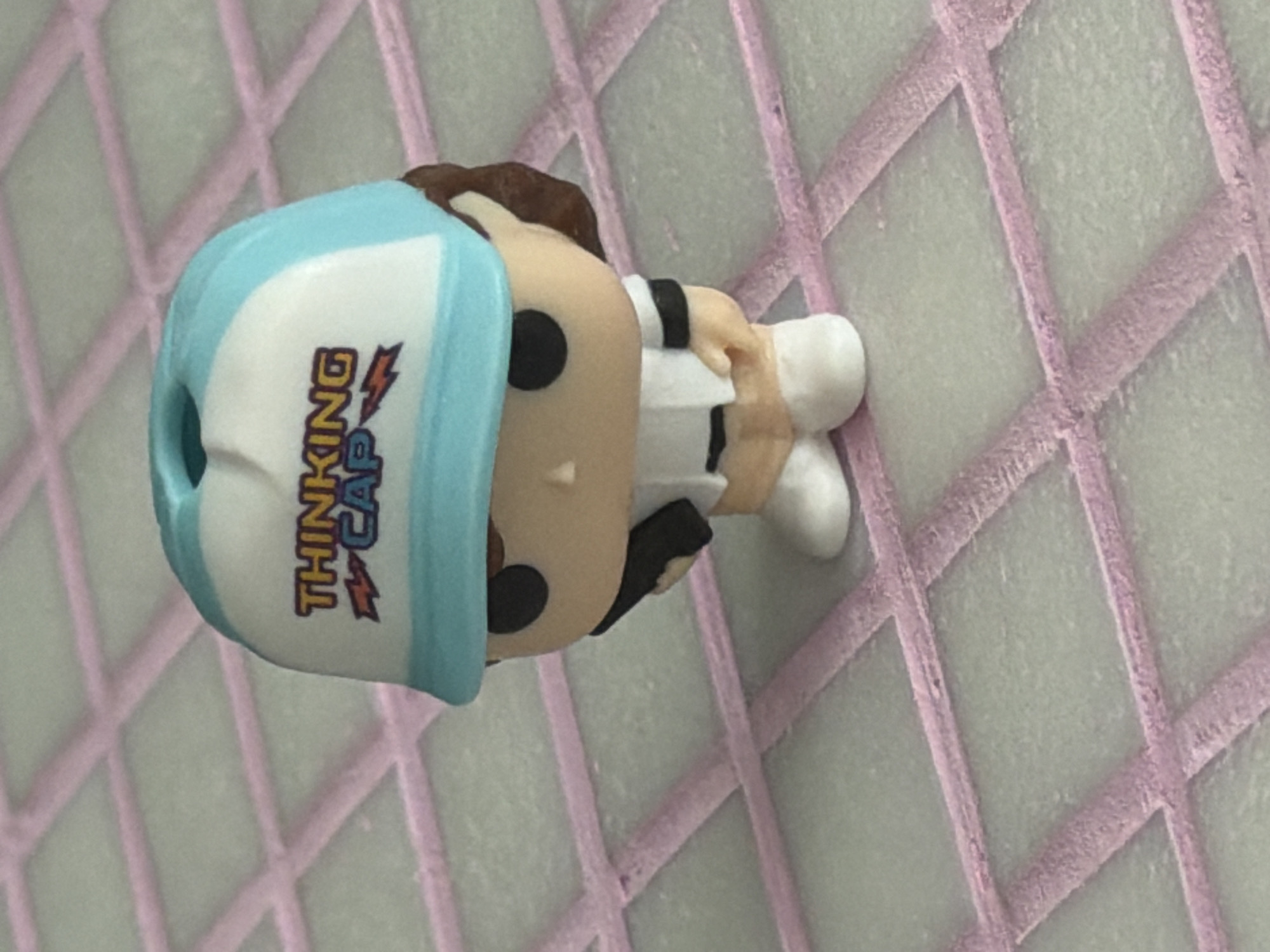 A miniature figurine of a person wearing a blue and white trucker hat with the words "THINKING CAP" and lightning bolt graphics is the sole subject of the image. The figurine has brown hair, black eyes, a light-skinned face, and is wearing a white shirt with a black stripe around the sleeve and light-colored shorts. It is holding a black object in its right hand. The figurine is standing on a surface with a grid pattern, consisting of light-colored squares outlined by pink grout. The background is out of focus and appears to be the same grid surface. The location is not discernible from the image.