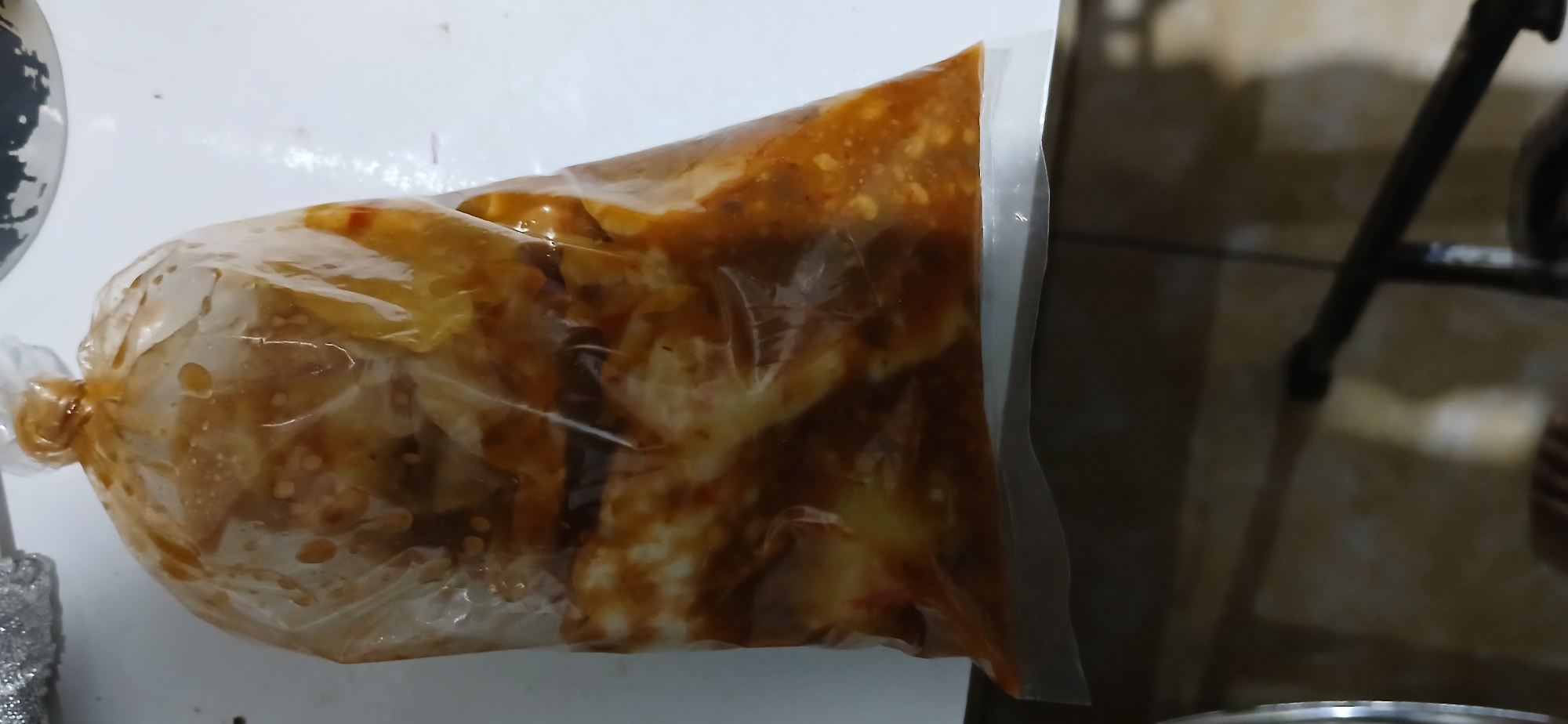 A sealed plastic bag containing food items coated in a reddish-brown sauce is positioned vertically against a white surface. The bag is tied at the top, creating a rounded shape. The food inside appears to be in chunks, possibly meat or vegetables, with a glossy coating from the sauce. The white surface extends to the left and right of the bag. Below the white surface, a dark, blurred background shows a tiled floor and a dark, horizontal object, possibly furniture or a piece of equipment, with a metallic sheen on the left side. The overall lighting is dim. The scene is not definitively identifiable as being in Cigombong, Indonesia.