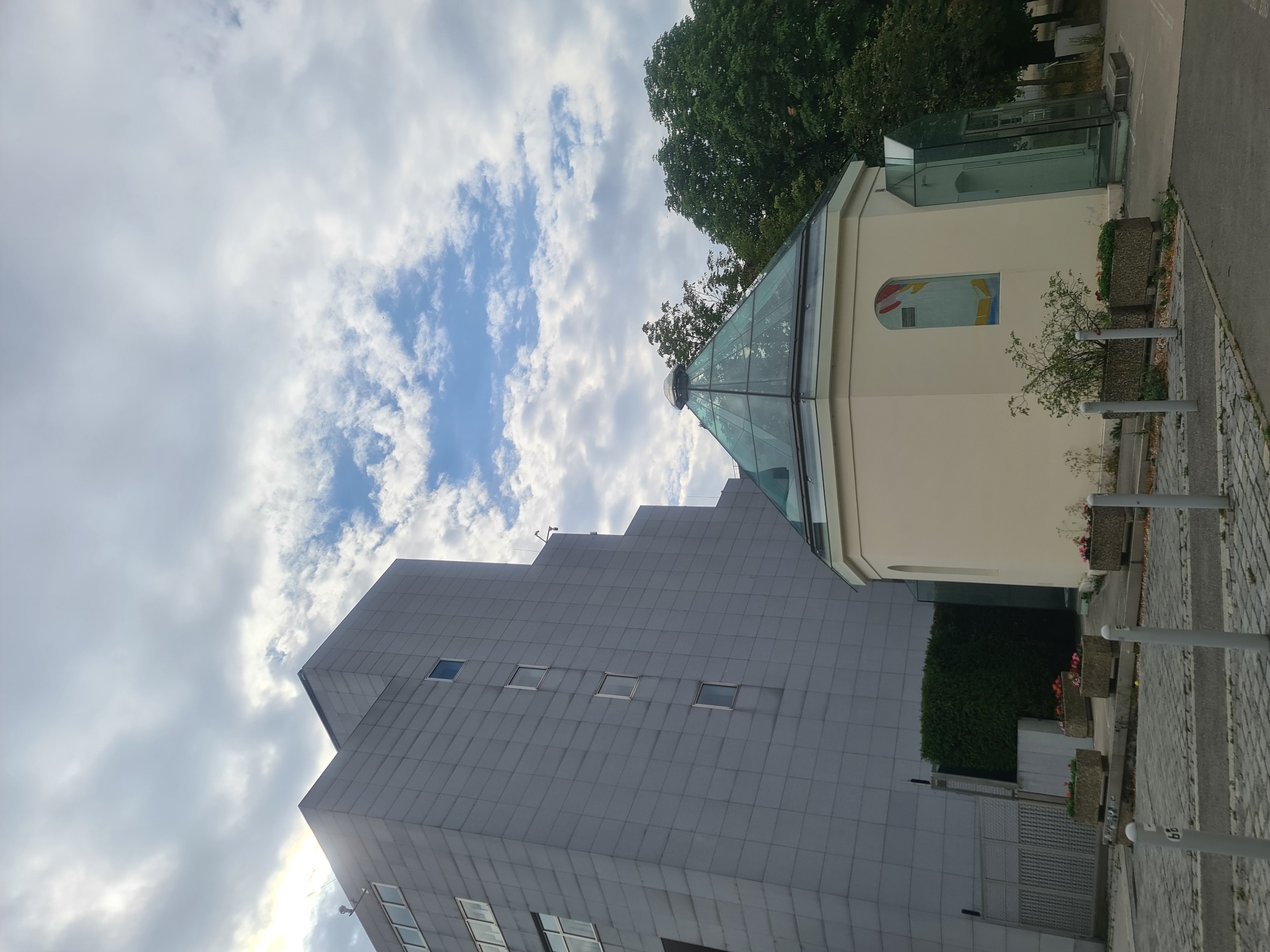 The image shows a modern building with a gray facade, composed of large rectangular panels. To the right of the modern building is a smaller, cream-colored octagonal structure topped with a glass dome. The octagonal structure has a stained-glass window with red and yellow elements, and a modern glass entrance. The scene is outdoors, with a cloudy sky and trees visible in the background. In the foreground, there is a cobblestone pathway, planters with flowers, and bollards. The location is identified as being in Vienna, Austria.