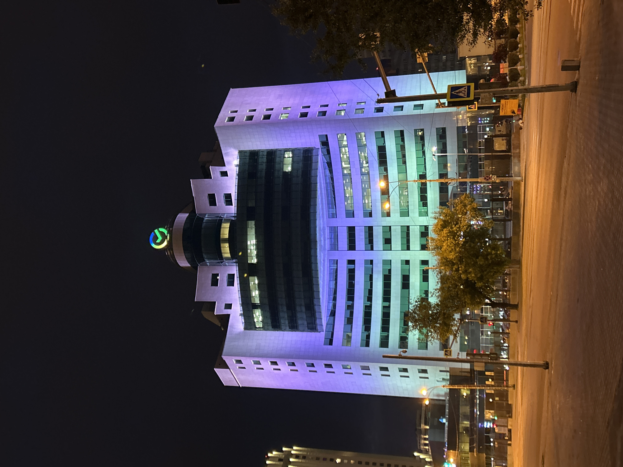 The image shows a nighttime view of the Sberbank building in Yekaterinburg, Russia. The building is illuminated with purple and green lights. A large Sberbank logo is visible at the top of the building. In the foreground, there is a street with lampposts and trees. There are also pedestrian crossings visible. The sky is dark, indicating it is nighttime. The scene appears to be a public space in a city.