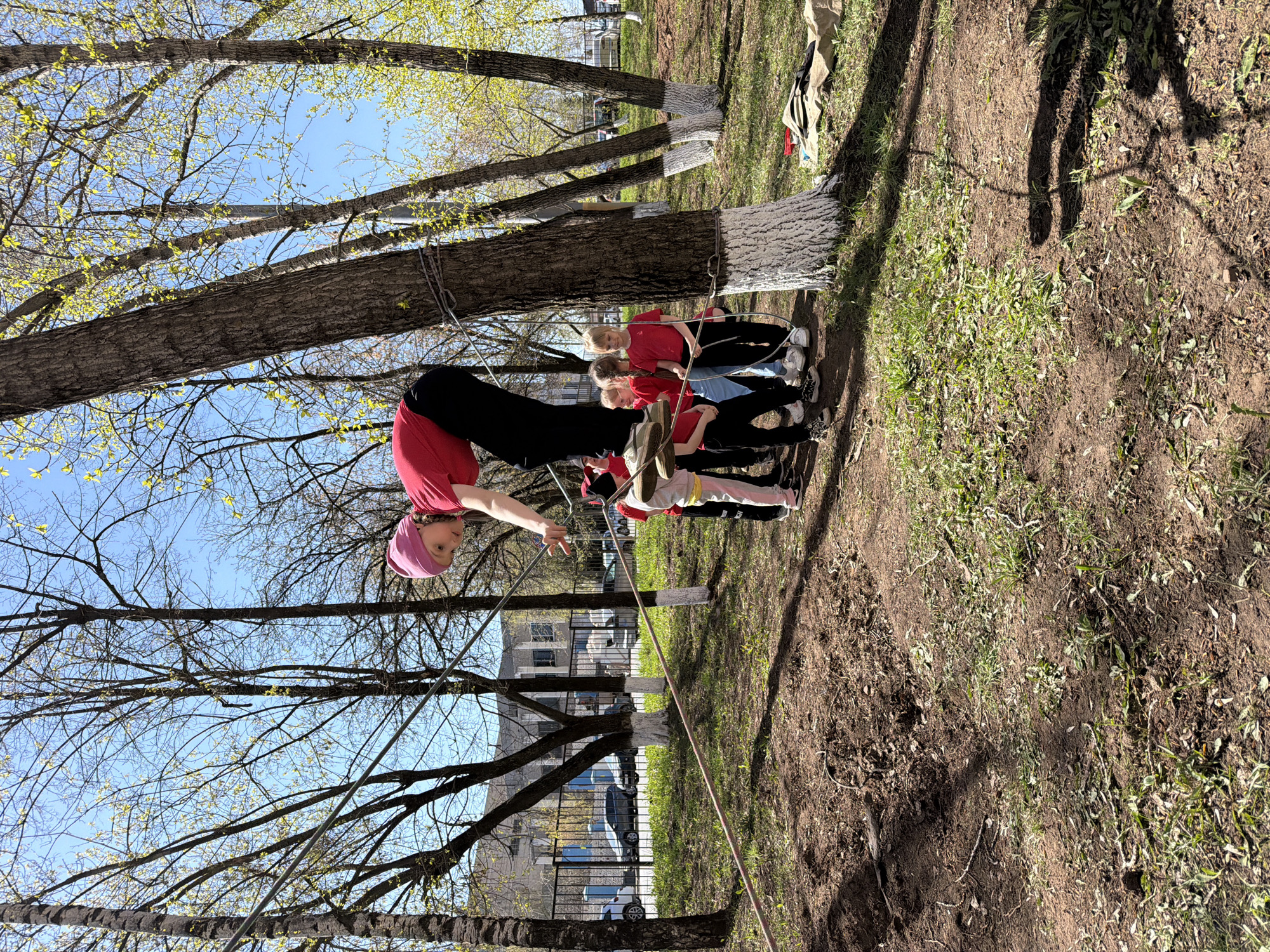 The image shows a group of people in what appears to be a park or grassy area in Izhevsk, Russia. The location is recognizable from the architecture in the background. One person, possibly a child or young adult, is suspended upside down, using a slackline or similar apparatus rigged between two trees. A group of other children or young adults are standing or kneeling nearby, seemingly observing or participating in the activity. The trees are partially bare, indicating it might be early spring or late autumn. The ground is a mix of dirt and grass. A rope or line is visible near the bottom of the frame, seemingly part of the setup for the suspended person. The sky is visible through the branches of the trees. The people are dressed in casual clothing.