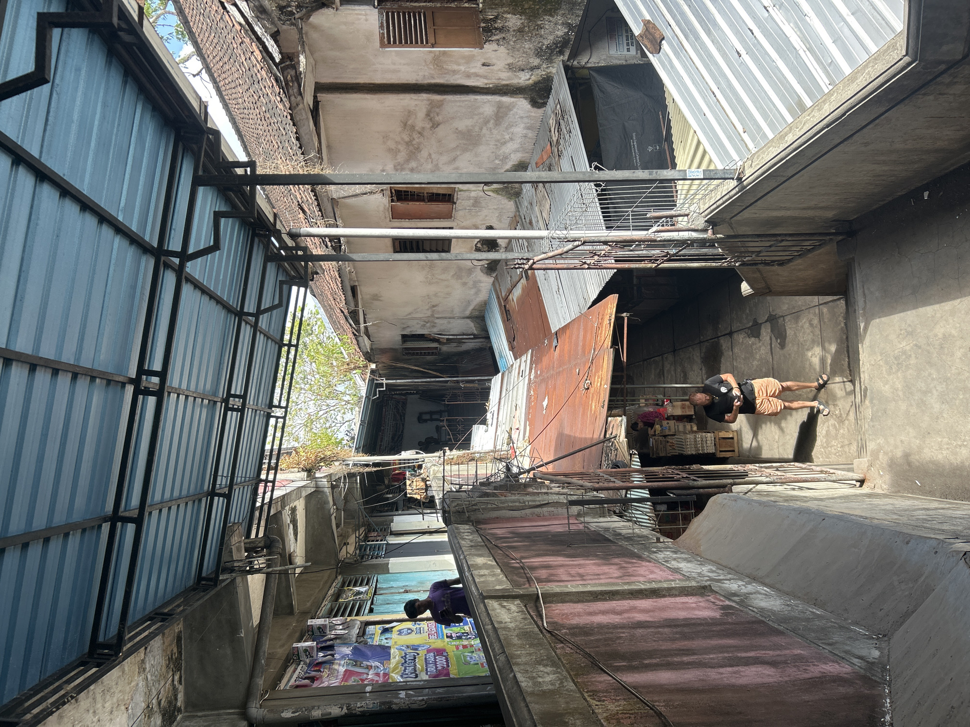 The image depicts a narrow alleyway in Denpasar, Indonesia, likely in a densely populated area. A blue corrugated metal roof with a black metal frame extends over the passageway. On the left, a multi-story building has a pinkish-red textured wall on its lower section and concrete on the upper section, adorned with various posters. A man in a purple t-shirt stands on an upper level, looking at his phone. The alleyway itself is paved with concrete. Further down the alley, a man in a black t-shirt and orange shorts is also looking at his phone. Another person, partially visible in a red and black patterned shirt, is in the background near stacked boxes. The buildings lining the alley are aged, with weathered walls and wooden shutters on the windows. Various corrugated metal roofs, some rusted, are visible on lower structures. The environment appears somewhat disorganized, with visible pipes and electrical wiring overhead. Sunlight casts shadows throughout the alley.