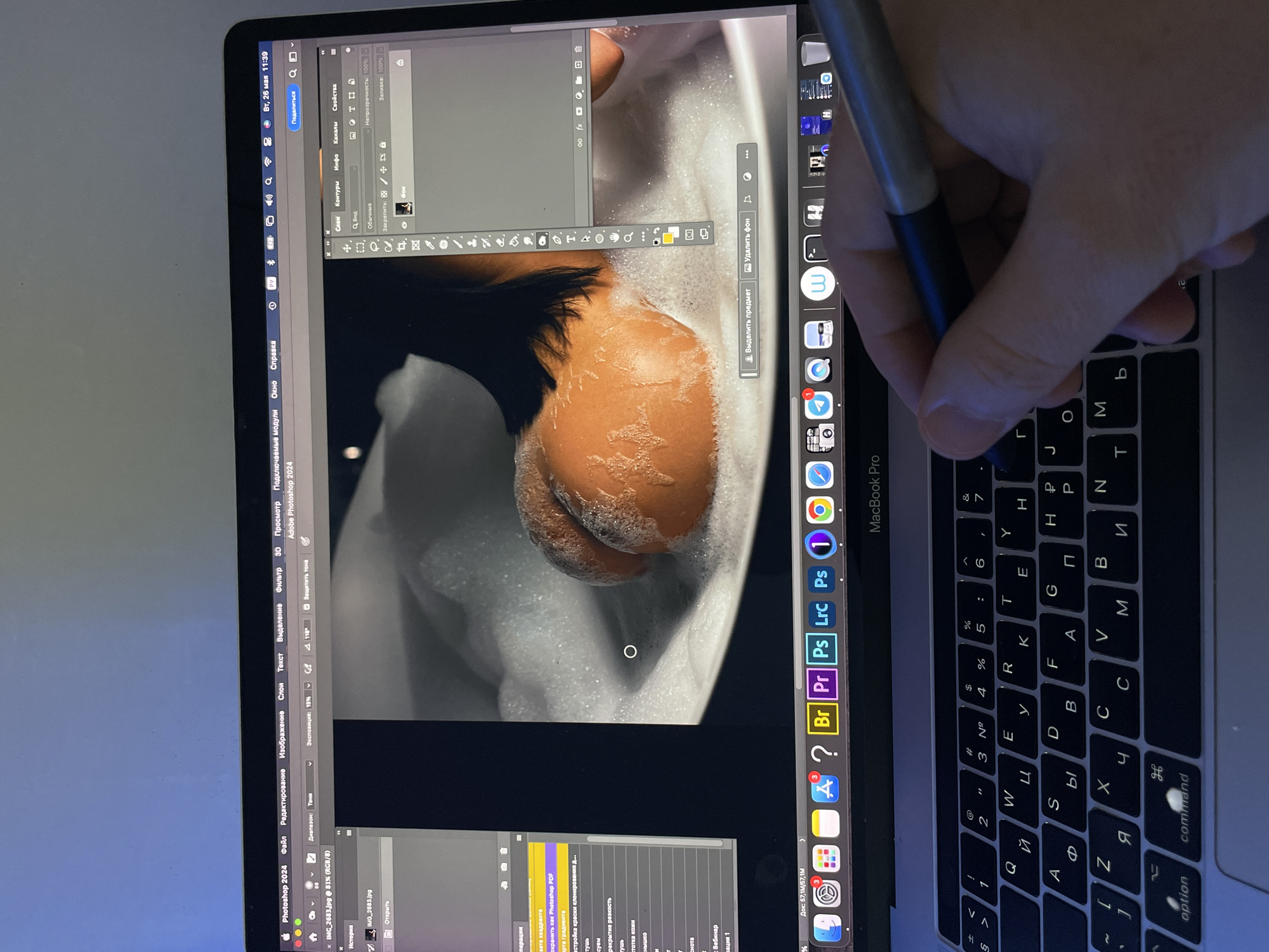 A person is using a stylus pen to edit a photo on a MacBook Pro, likely in Adobe Photoshop 2024. The photo being edited shows a dark-haired person in a bathtub filled with bubbles. The keyboard of the laptop is visible, with Russian letters alongside English. The laptop's dock displays various applications including Adobe products. The date and time are shown as Tuesday, May 26, 11:39. The location is likely Kazan, Russia, indicated by the Russian keyboard layout.