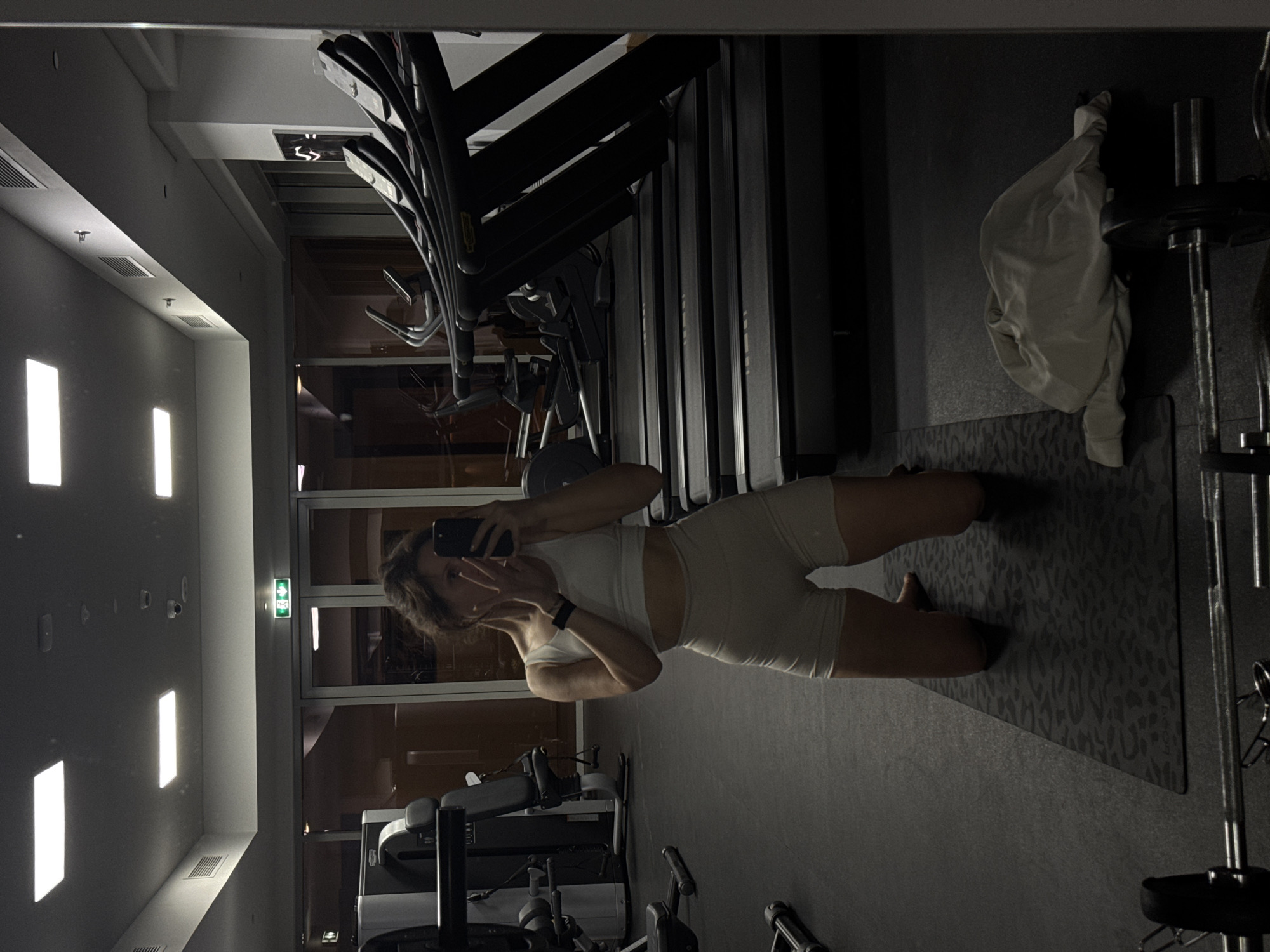 The image shows a woman taking a selfie in what appears to be a gym. She is wearing a light-colored athletic outfit consisting of a sports bra and shorts. She is kneeling on a patterned yoga mat. Behind her, there are several treadmills lined up, and weightlifting equipment is visible to her right. The setting seems to be indoors, with large windows in the background reflecting the interior lights, potentially at night. Based on the image details alone, there's no clear indication or evidence to confirm its location in Dubai, United Arab Emirates. More information or contextual cues from the image would be necessary for such verification.