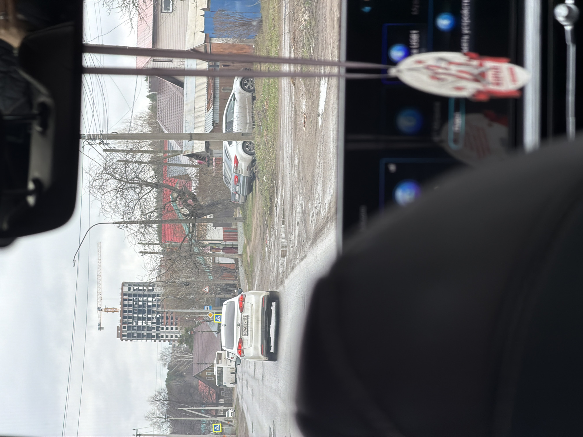 This media file captures a street scene in Chaikovsky, Russia, viewed from inside a vehicle. The image is taken from a side window, looking out onto a wet, likely muddy, road under an overcast sky.
In the immediate foreground, a dark car interior is visible on the left, partially obscuring the view. A small, white and red, spaceship-like ornament hangs near a blurry digital screen on the right, which displays blue icons and some indistinct Cyrillic text.
Outside the vehicle, a white SUV is visible from the rear, traveling away from the viewer. Its license plate, "H940EK159," clearly displays the region code "159," which corresponds to Perm Krai, Russia. Further down the street, other cars are parked or driving. The road surface appears damp and uneven, suggesting recent rain or unpaved sections.
On the right side of the road, a row of houses with varied architecture and roof colors (brown, red, blue) lines the street. Bare trees, indicative of late autumn, winter, or early spring, are interspersed among the buildings. Utility poles with numerous wires are also visible.
To the left of the road, a large construction site is prominent, featuring a partially built multi-story building constructed of concrete, with many empty window frames. A tall yellow construction crane rises above the unfinished structure against the cloudy sky. Pedestrian crossing signs are visible along the roadside. The overall scene depicts a typical, somewhat dreary, daytime setting in a Russian town.