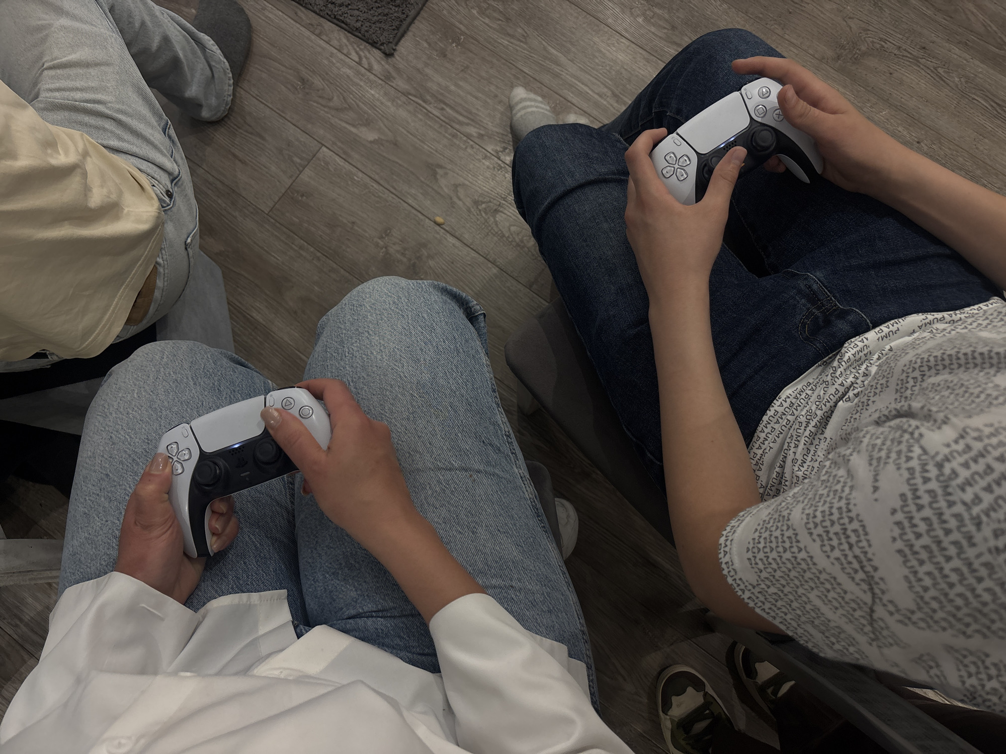 An overhead view captures two individuals engaged in a video game session, likely indoors in Yerevan, Armenia. Both are seated, with their hands holding white PlayStation 5 (PS5) controllers.
The person on the left is wearing light-wash blue jeans and a white long-sleeved shirt. Their hands grip a PS5 controller, which shows a subtle blue light from its central touch bar. Their fingernails are painted a light, neutral color. Another person's leg, also in light-wash jeans, is partially visible further up the frame.
The person on the right is dressed in dark-wash blue jeans and a white short-sleeved shirt. The shirt is distinctly patterned with the repeated text "PUMA". They are also holding a white PS5 controller. A grey striped sock is visible on their left foot, and a camouflage-patterned sneaker on their right foot.
The setting features a light grey wooden floor, with a small dark grey rug visible in the background. The scene conveys a relaxed, casual atmosphere centered around shared entertainment.