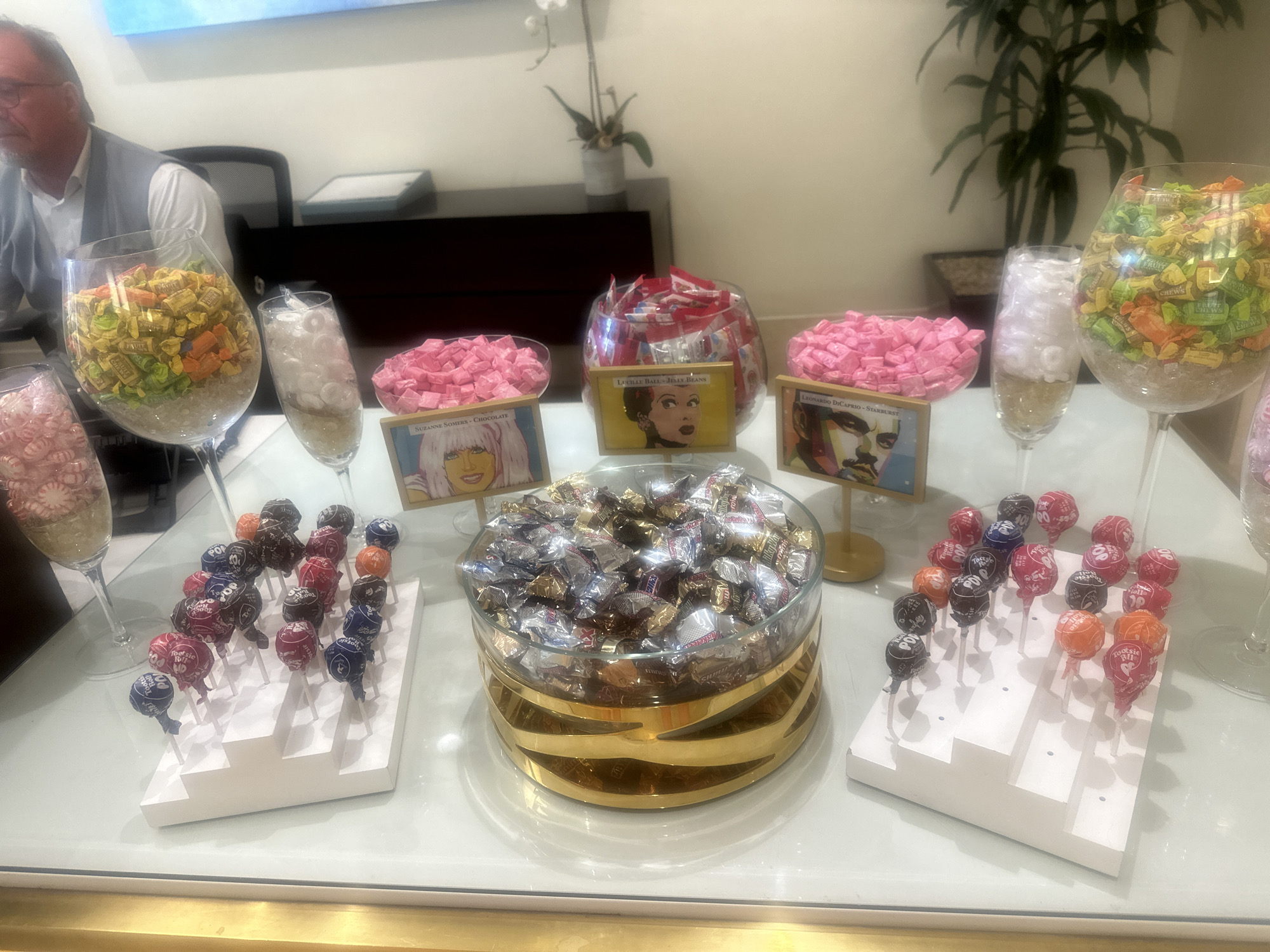 A table with an assortment of candies is shown in this medium shot. A man is visible on the left, wearing a white shirt and gray vest. Behind him is a black office chair.
Centered on the table are several glass containers filled with different types of candy. From left to right: a tall glass filled with pink and white peppermints, a tall glass filled with yellow and orange fruit chews, a tall glass filled with white candies, a bowl filled with pink Starbursts, a bowl filled with red candies, a bowl filled with pink Starbursts, a tall glass filled with white candies, and a tall glass filled with yellow and orange fruit chews. Between the candy bowls are signs featuring images of celebrities like Suzanne Somers, Lucille Ball, and Leonardo DiCaprio.
In front of the center row of candy are two stands of lollipops in assorted colors, and in the middle of them, a glass bowl with a gold base filled with chocolate candies wrapped in silver foil.
A green plant can be seen in the upper right of the image. The location is Unknown, Unknown.