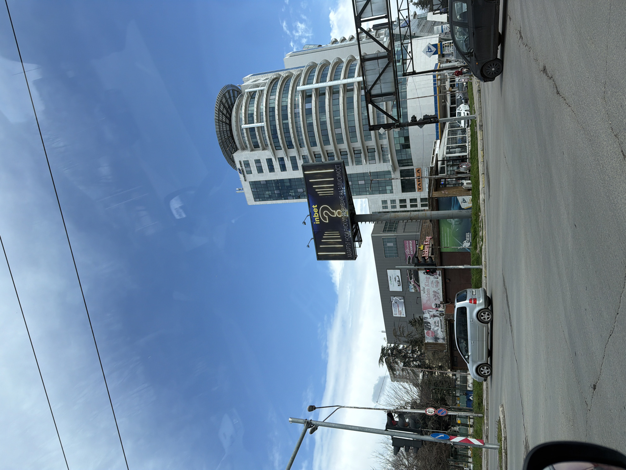 The image depicts an urban street scene at the intersection of Osmi Primorski Polk Boulevard and Hristo Botev Boulevard in Varna, Bulgaria, under a partly cloudy blue sky. The viewpoint is from within a vehicle, with a portion of a rearview mirror visible in the lower-left foreground.
The asphalt roadway displays cracks and general wear. A silver minivan, consistent with a Volkswagen Touran, is positioned in the left lane, moving from left to right across the frame. A dark-colored hatchback is partially visible in the right lane, facing towards the viewer.
On the left, a metal utility pole supports a traffic light, illuminated red, and a street lamp. Below the traffic light, regulatory signs include a circular '40' speed limit sign and a blue circular sign with a white arrow pointing to the right. Bare-branched trees line the sidewalk on the left.
The midground features various commercial buildings and infrastructure. A large, illuminated billboard with "inbet ?" and the Bulgarian text "ХАЗАРТЪТ НОСИ РИСК ОТ РАЗВИВАНЕ НА ЗАВИСИМОСТ" (Gambling carries a risk of developing addiction) stands prominently. Directly behind it, the multi-story Energo-Pro Business Center, a modern white building with both rectangular and curved architectural sections, features numerous windows and a helipad structure on its roof. The text "ENERGO-PRO" is affixed to its upper facade.
To the left of the Energo-Pro center, a lower, grey commercial building displays multiple advertisements, including "Chicken King," "MA AutoBox," and "DIESEL." Adjacent to the roadway on the right, a "TABLA" restaurant or cafe with a glass enclosure is visible, situated next to a "Petrol" branded gas station. One individual wearing a red top is observed standing near the "Petrol" station. Overhead utility wires traverse the upper portion of the sky.