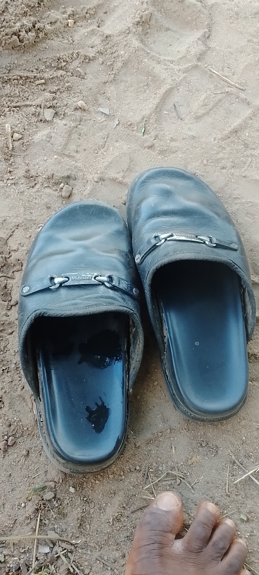 The image shows a pair of black, well-worn "Minister" brand slip-on sandals resting on sandy soil in Adankolo, Nigeria. A dark-skinned foot is partially visible in the lower right corner of the frame. The ground is light brown and shows evidence of vehicle tire tracks.