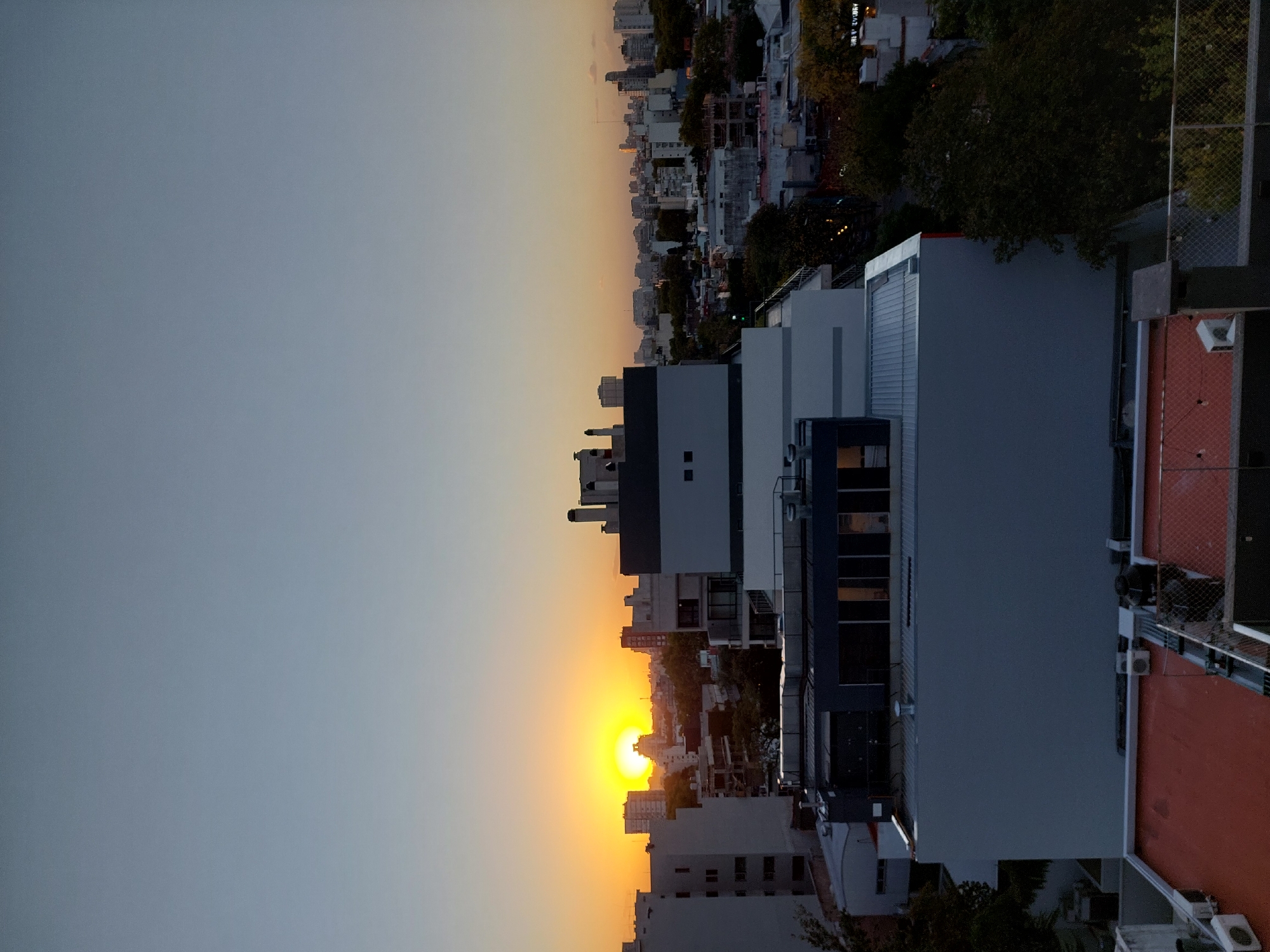 The image captures a sunset view over the city of Buenos Aires, Argentina. The sky transitions from a pale blue to a vibrant orange where the sun is setting on the horizon. In the foreground are various buildings and rooftops, including a modern multi-story building with a white facade and dark blue accents. A chain-link fence and some rooftop equipment are visible. Beyond the immediate buildings, the urban landscape stretches out, with a dense collection of buildings of varying heights and architectural styles. Some trees dot the cityscape, providing patches of green. The overall impression is an urban sunset scene.