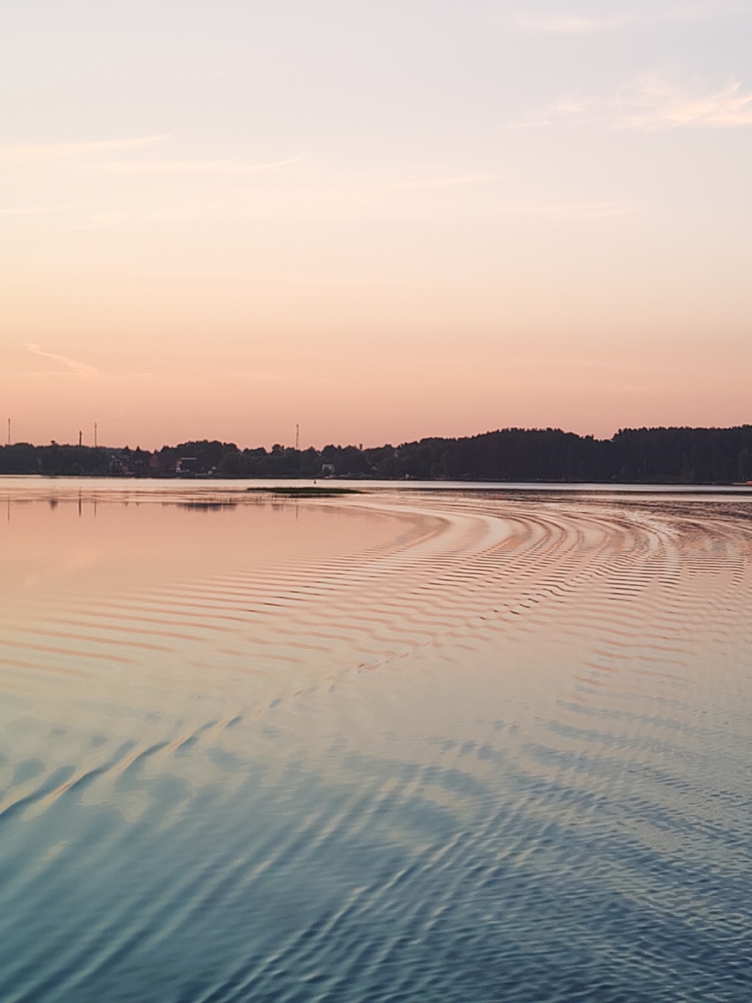 The image shows a tranquil body of water reflecting the soft colors of a sunset. The sky is a gradient of pale pink and light blue, with a few wispy clouds. On the horizon, a dark line of trees and some distant buildings with tall, slender structures that resemble communication towers are visible. The water's surface is disturbed by gentle ripples, creating a pattern of light and shadow that extends from the right side of the frame. The overall impression is one of peacefulness and natural beauty. The location is identified as Terekhovo, Russia.