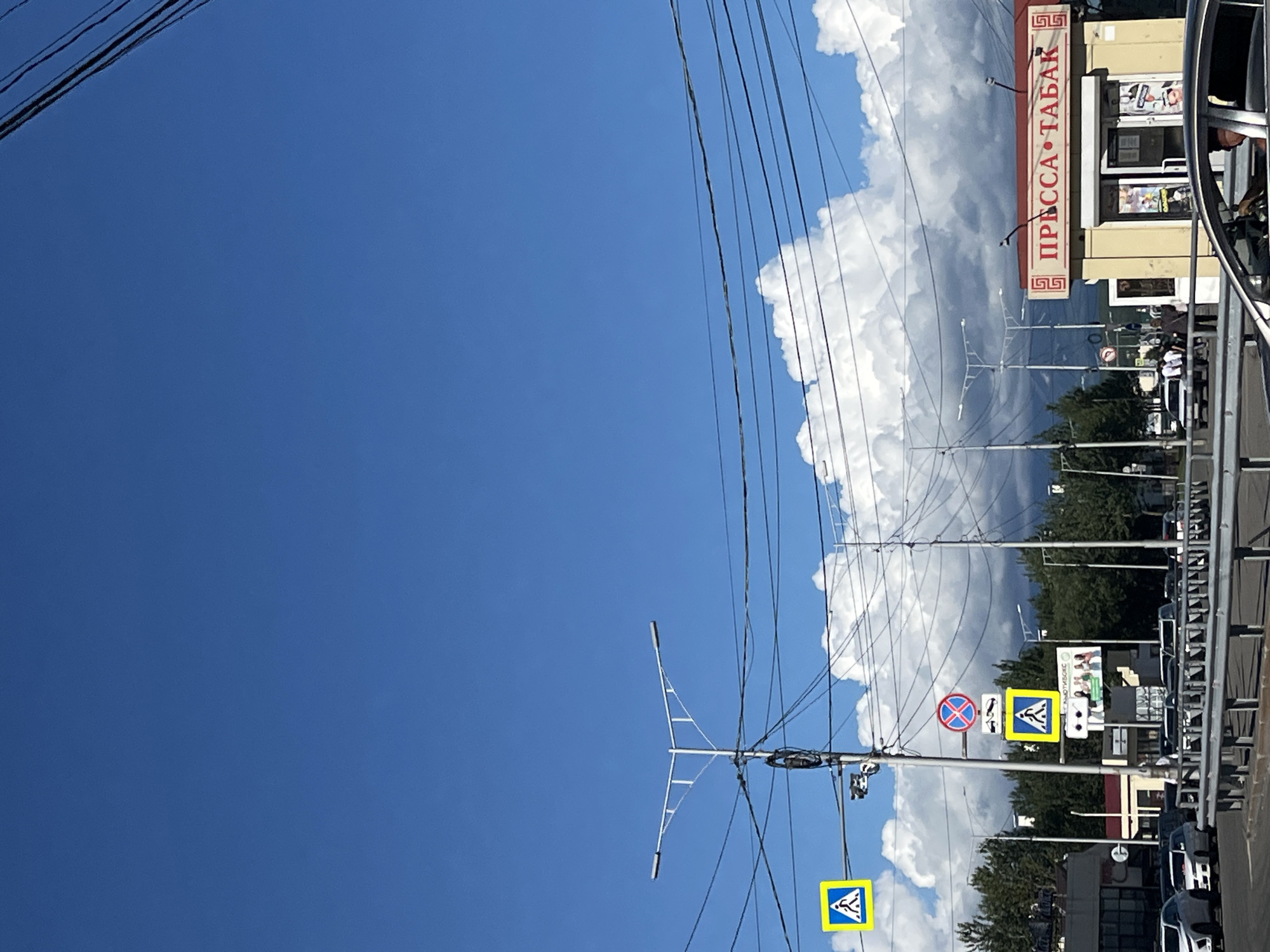 The image depicts a street scene in Petropavlovsk-Kamchatsky, Russia, under a bright blue sky with scattered white clouds. A pedestrian crossing signal with a yellow diamond-shaped sign indicating a pedestrian crossing is visible on the left. Above it, a street lamp with two lights extends outwards. Further down the street, another pedestrian crossing sign is present, accompanied by a circular red and blue traffic sign indicating no stopping.
In the middle ground, several cars are parked and moving. A kiosk with a red roof, labeled in Russian as "ПРЕССА • ТАБАК" (Pressa • Tabak, meaning "Newspaper • Tobacco"), is situated on the right side of the image. The kiosk has a glass storefront displaying various items. Several streetlights are scattered across the background, and a network of overhead power lines traverses the upper portion of the image. Trees and buildings can be seen behind the street and kiosk. In the foreground, a section of a metal guardrail separates the road from the viewer. A car with a driver is partially visible in the lower right corner.