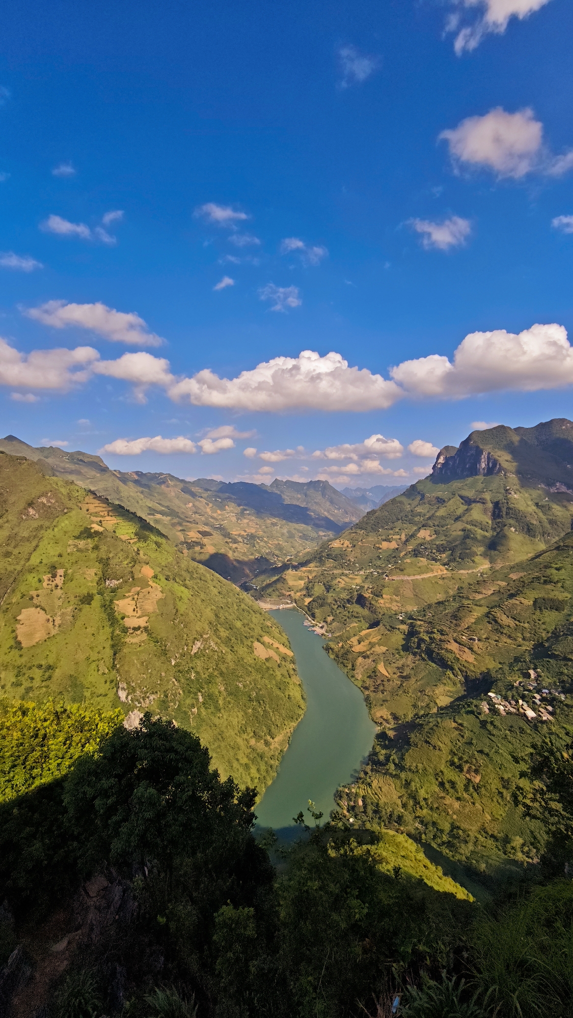 The image shows a deep river valley in the Đồng Văn Commune, Vietnam, as viewed from a high vantage point. Lush green hillsides with terraced fields dominate the landscape on either side of the river. The river itself is a pale green color and winds through the valley. The sky is a brilliant blue with scattered puffy white clouds. The foreground of the image contains dark green foliage. Distant mountains can be seen in the background.