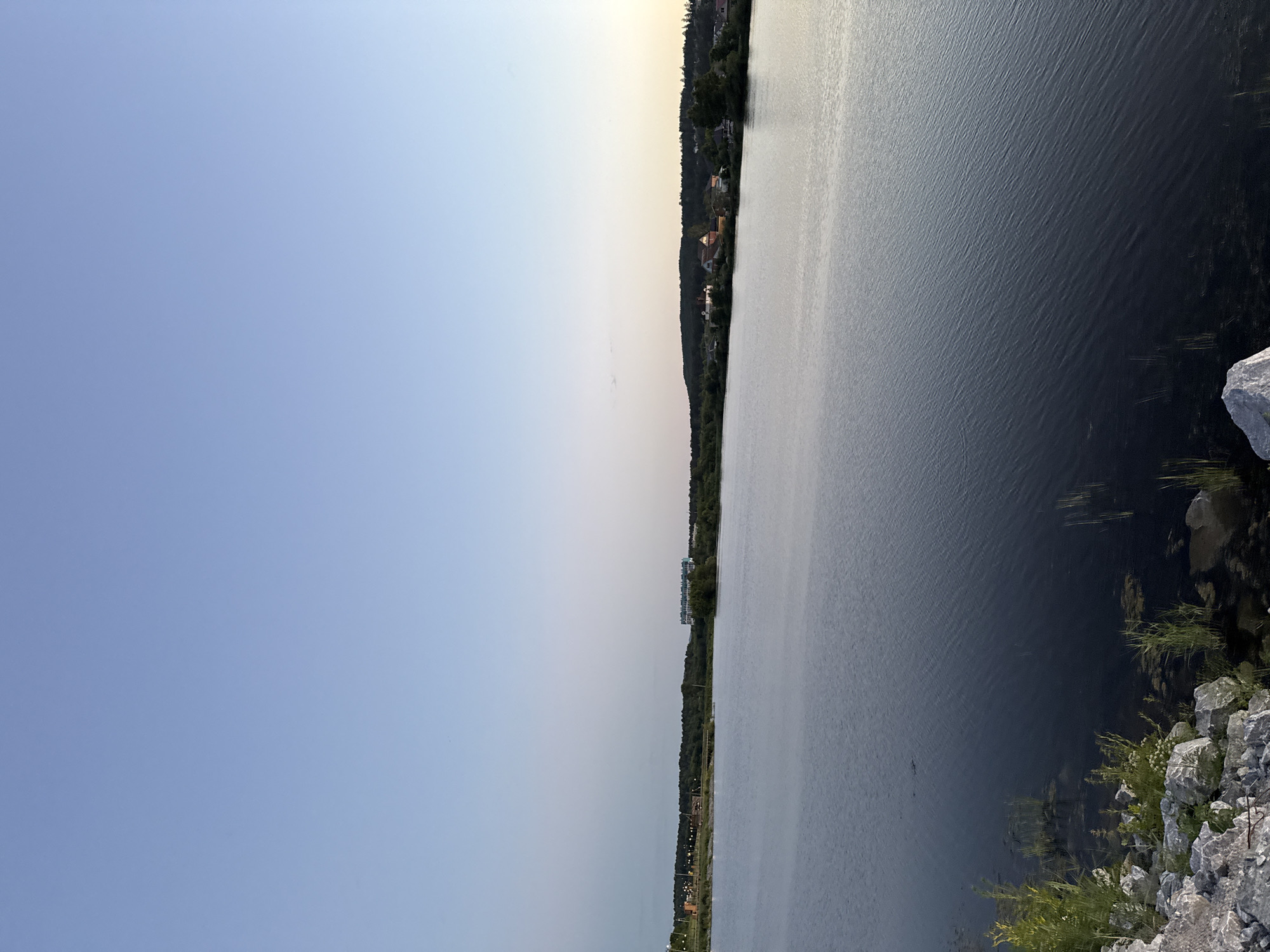 The image depicts a wide, calm body of water, likely a lake or reservoir, under a clear, pale blue sky transitioning to a lighter hue near the horizon. The foreground shows a rocky shoreline with sparse green vegetation and reeds growing in the shallow water. In the mid-ground and background, a tree-lined horizon is visible, with a few structures, including what appears to be a residential area with houses and a larger, modern building with light-colored facades, suggesting an urban or suburban fringe. The lighting indicates it is either early morning or late evening, given the soft, diffused light and the clear, cloudless sky. The scene is tranquil and natural, with hints of human development on the distant shore. The location is identified as the Turgoyak Lake in Miass, Russia, a well-known natural landmark.