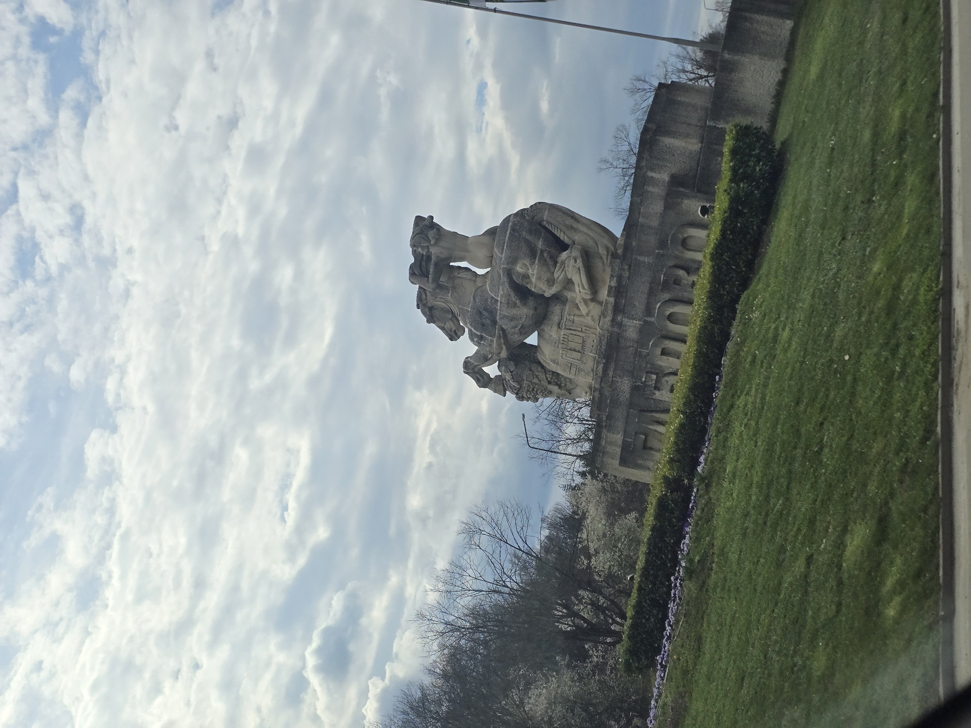 The image shows a statue in Gabrovo, Bulgaria, depicting a figure atop a horse. The statue is located on a grassy area beside a road. The sky is cloudy. There are trees and bushes around the statue.