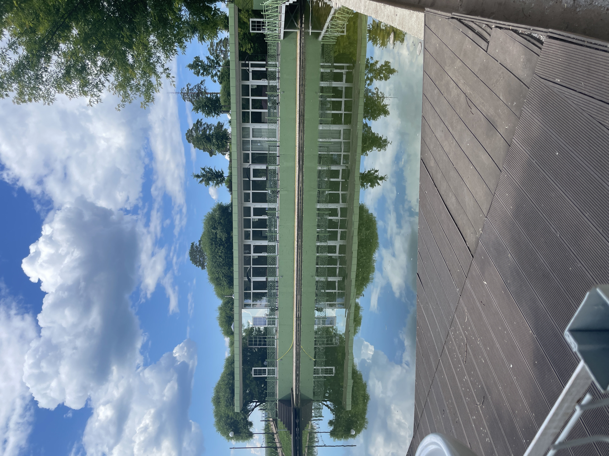 The image depicts a modern green building with large windows, situated alongside a calm body of water. The building's facade is reflected in the water, creating a symmetrical visual effect. Lush green trees and a bright blue sky with fluffy white clouds form the backdrop. In the foreground, a wooden deck extends towards the water, partially visible on the lower right and left. The scene appears to be a tranquil outdoor setting, possibly a park or a waterfront recreational area. The specific location in Izhevsk, Russia, is not discernible from the visual information alone, however, such a structure might be a restaurant, cafe, or a public building within a park or resort area in Izhevsk, Russia.