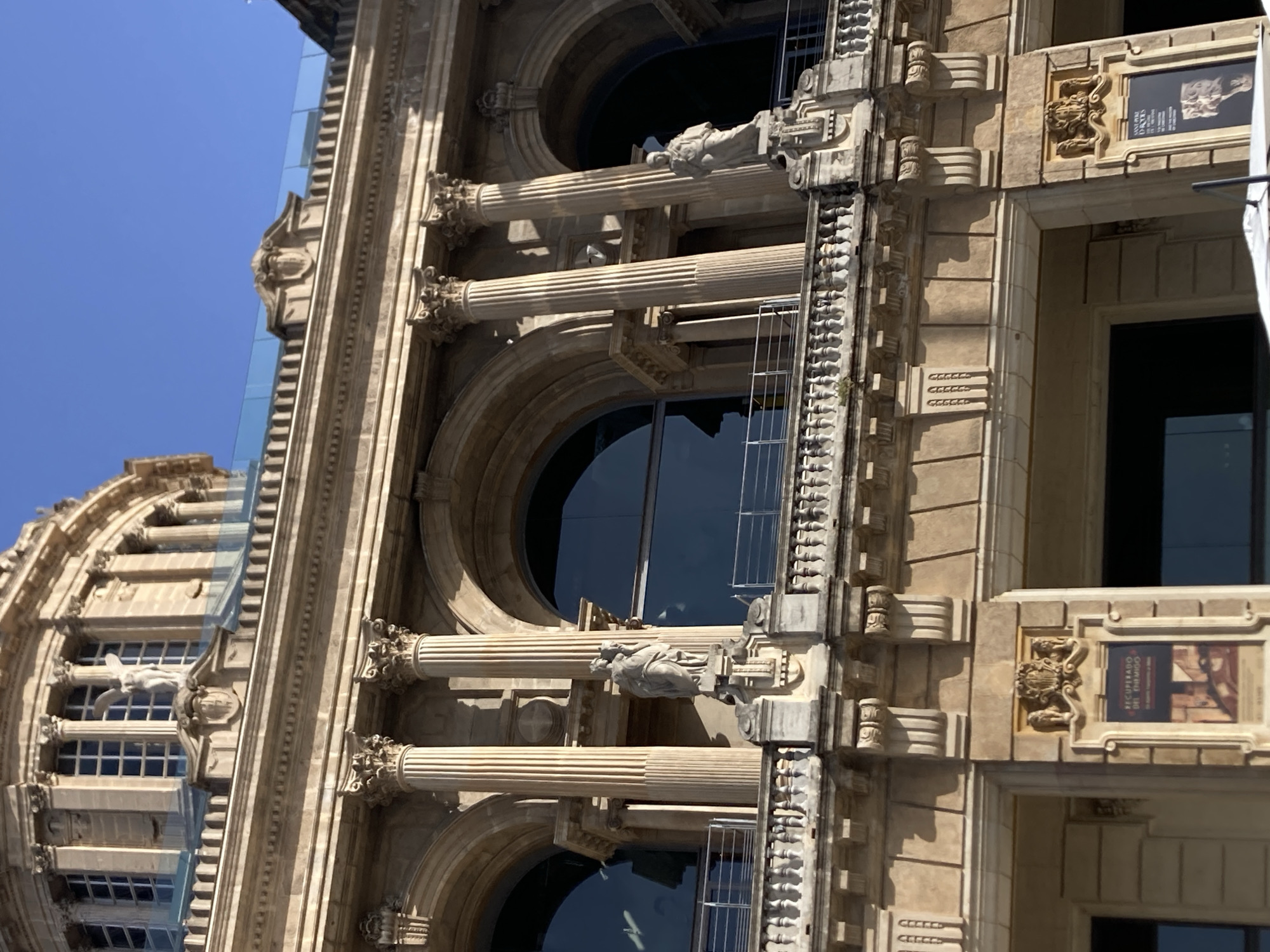 The image shows a close-up of a section of the Palau Nacional in Barcelona, Spain, on a clear day. The architecture is classical, with large arched windows, Corinthian columns, and ornate stone carvings. Two sculptures of human figures are visible on a balcony, one on the left and one on the right. The sculpture on the left depicts two figures embracing, while the one on the right shows a single standing figure. There are also decorative moldings and a balustrade along the balcony. Below the balcony, posters for exhibitions are visible on the building's facade. The sky is a vibrant blue, and sunlight casts shadows on the building's intricate details.