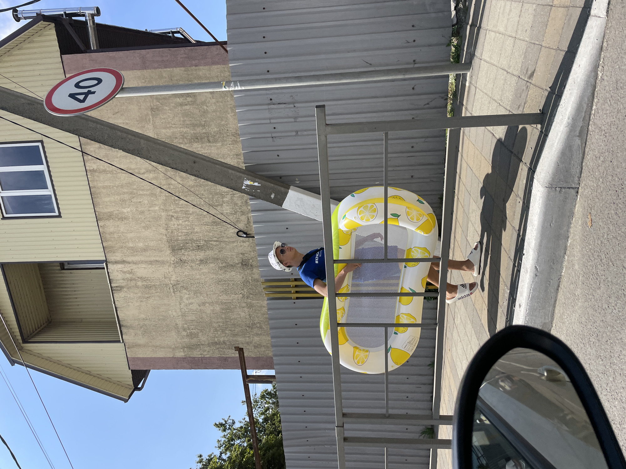 A young person wearing a white bucket hat, sunglasses, a blue t-shirt, and white slide sandals walks along a sidewalk carrying a yellow and white lemon-patterned inflatable pool float. The person is walking past a grey corrugated metal fence and a metal railing. In the background, a building with a beige facade and a yellow-paneled upper section is visible, along with a speed limit sign indicating 40. The scene appears to be in Dzhubga, Russia, given the context. The sun is shining, casting shadows on the ground, and the sky is clear and blue. A car's side mirror is visible in the lower left corner of the frame.