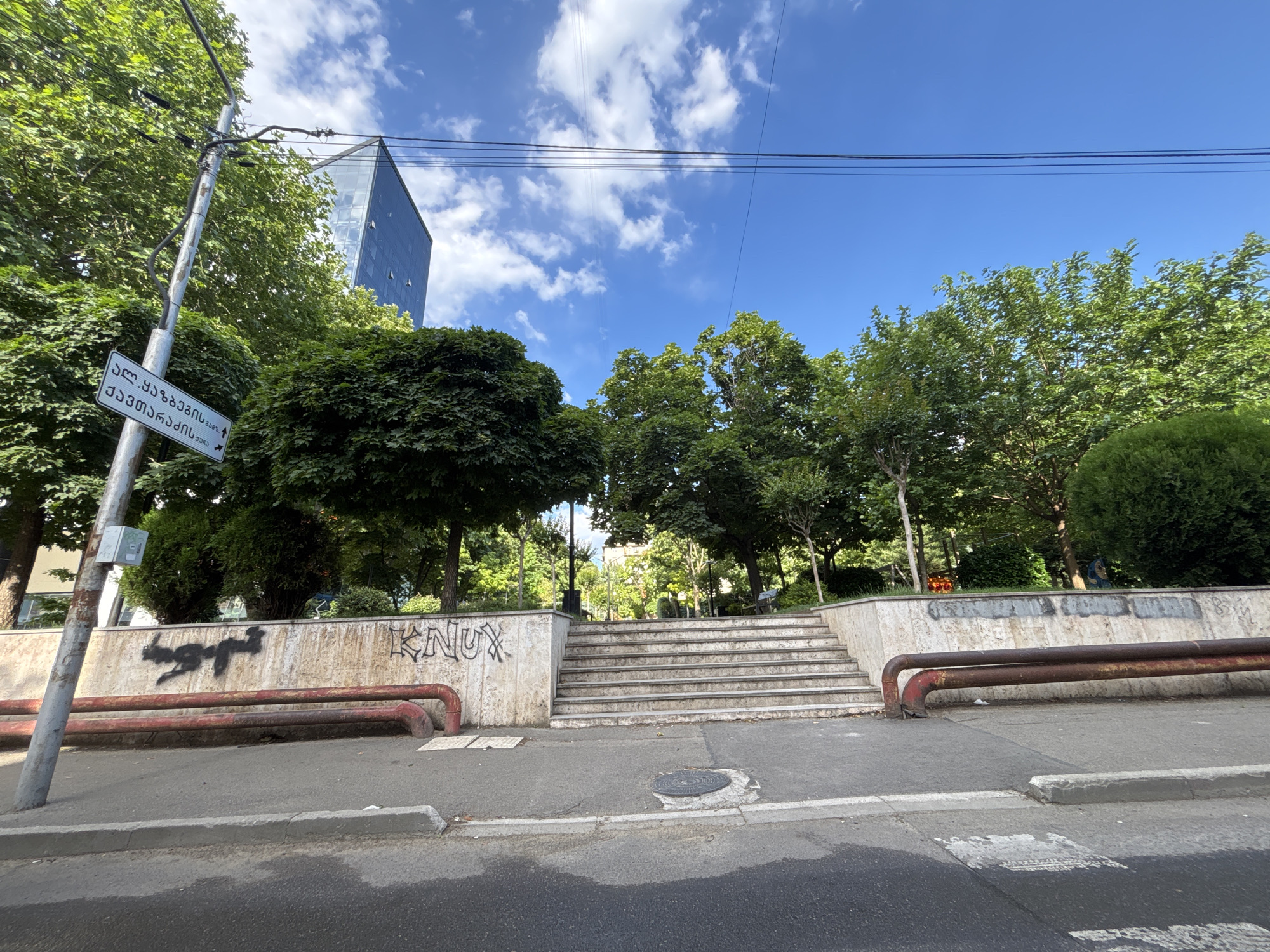 The image captures a view of a street in Tbilisi, Georgia, featuring a set of stone stairs leading up to a park or elevated area. The stairs are flanked by concrete walls adorned with graffiti, including the word "KNU" and an abstract spray-painted figure. Two rusted metal pipes run horizontally along the base of the wall. A street sign written in Georgian script is visible on a lamppost to the left, pointing towards the direction of the stairs. Lush green trees and a modern glass-fronted building rise in the background against a bright blue sky with scattered white clouds. The foreground shows a paved street with a visible curb and a manhole cover.