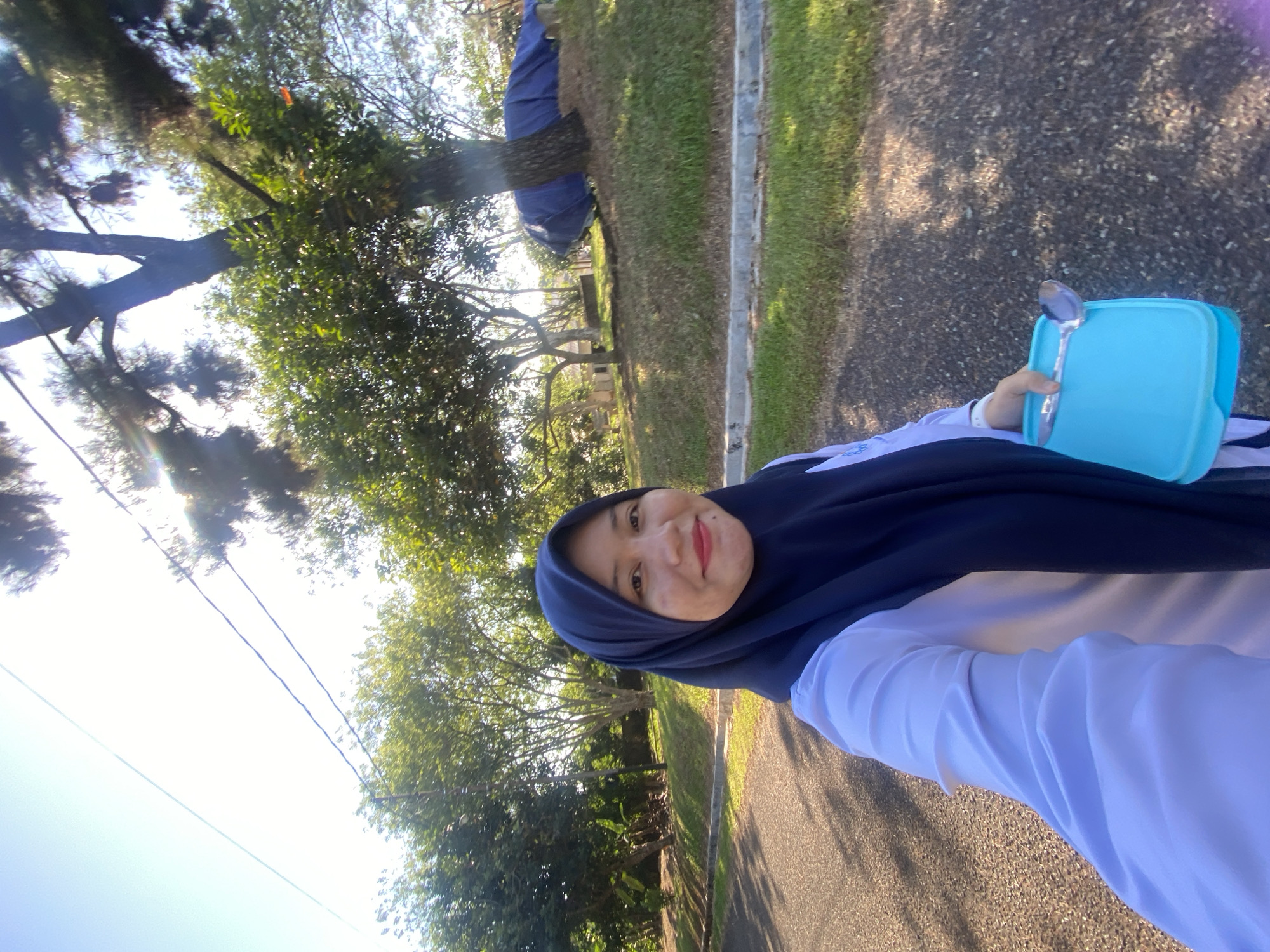 A woman wearing a navy blue hijab and a white long-sleeved shirt is taking a selfie outdoors. She is holding a light blue lunchbox with a spoon in her right hand. The background consists of a paved road, a grassy incline, and several trees. A blue tarp is visible covering something on the grassy slope. The scene appears to be in a park or residential area in Banjarbaru, Indonesia, as indicated by the presence of tropical vegetation and typical Indonesian road infrastructure. The sun is shining brightly, casting shadows.