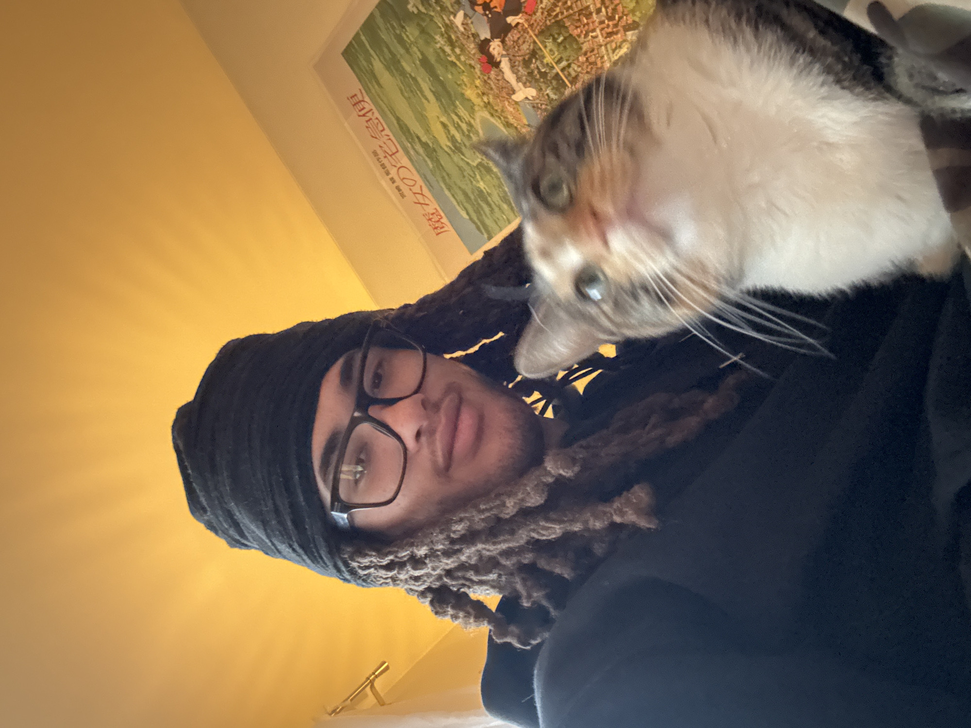 A selfie shows a man with long, tan dreadlocks, wearing a black head covering and dark-framed glasses, looking at the camera. A cat with white, gray, and light brown fur is held up near his face. The background includes a light yellow wall, a poster, and part of a dark-colored shirt. The location and specific details about Paris, France are not visible in the image.