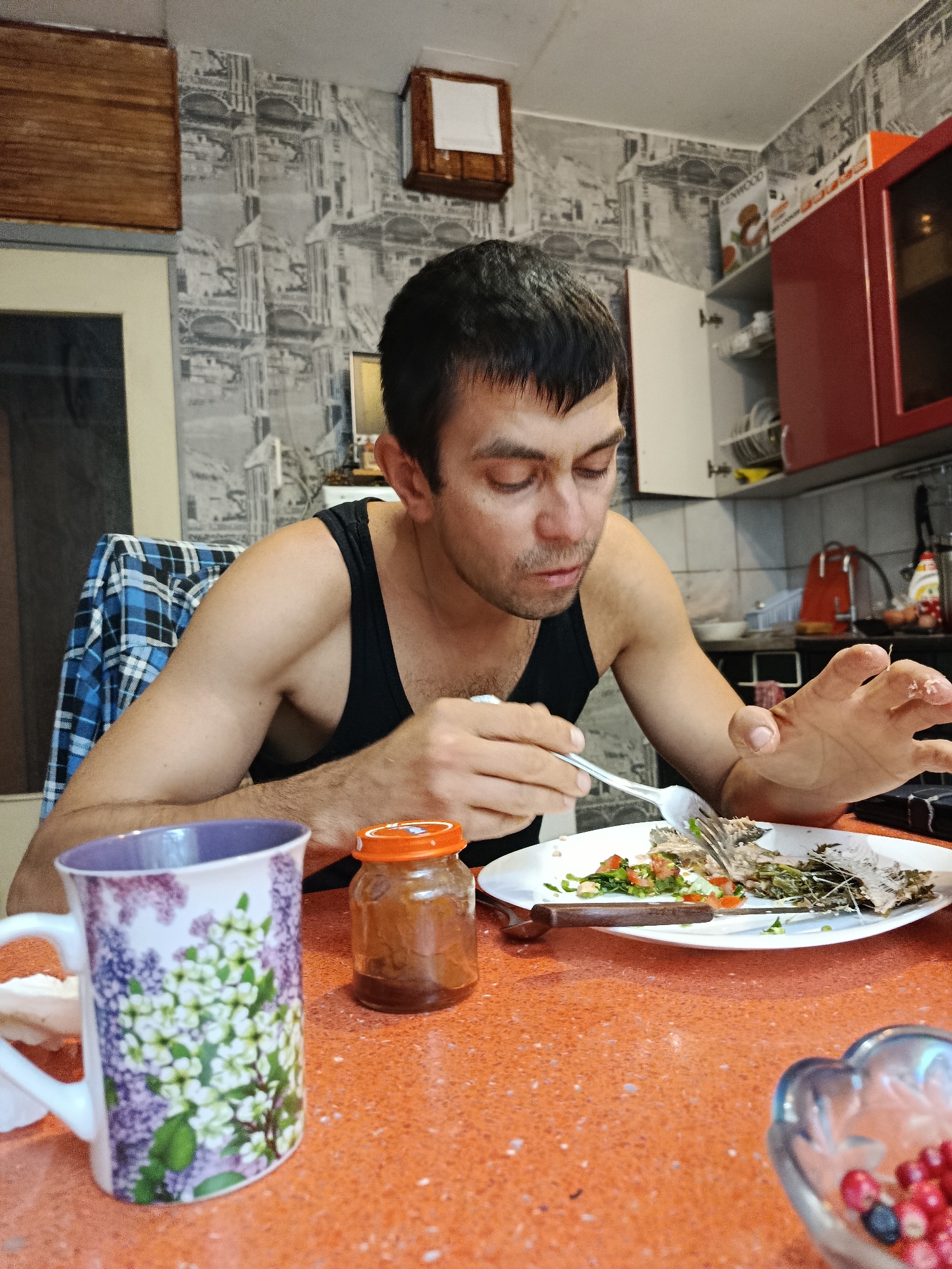 A man with dark hair and a thin build is seated at a bright orange table, eating fish from a white plate. He wears a black tank top and is using a fork to eat. To his left is a mug with a floral pattern, and a small glass jar with an orange lid. In the background, a kitchen is visible, with red cabinets and a patterned wallpaper. A blue and white plaid shirt is draped over the back of the man's chair.