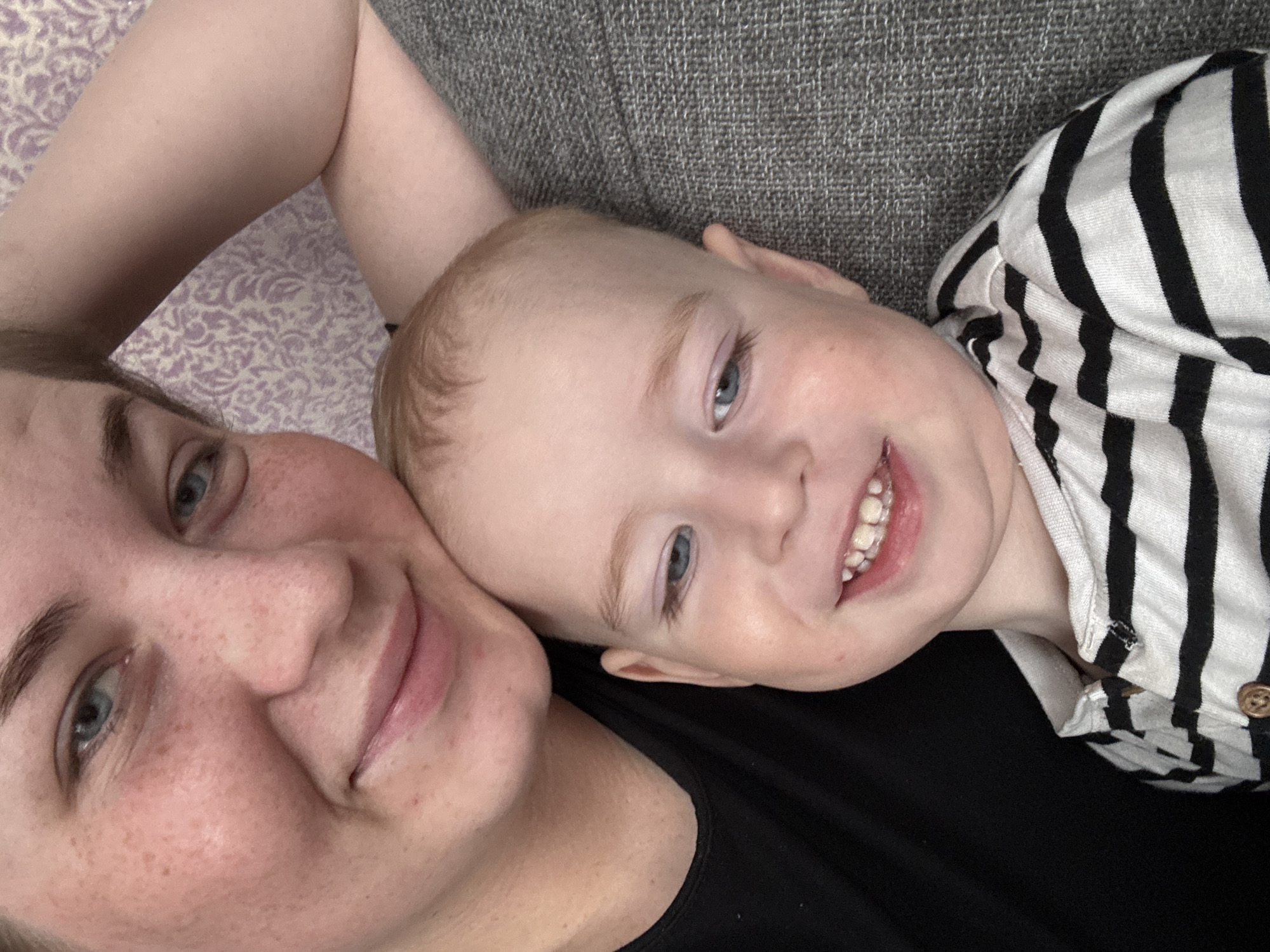 The image shows a selfie of a fair-skinned woman and a young child. The woman has light eyes, freckles, and is wearing a black top. The child is fair-skinned with light hair and light eyes, wearing a striped shirt with black stripes on a white background. The child is smiling, and the woman appears to be smiling slightly as well. They are likely lying on a couch or bed, with a gray textured surface visible behind the child and a patterned surface behind the woman's arm. The location in the city of Уральское, Russia, is not evident from the image.