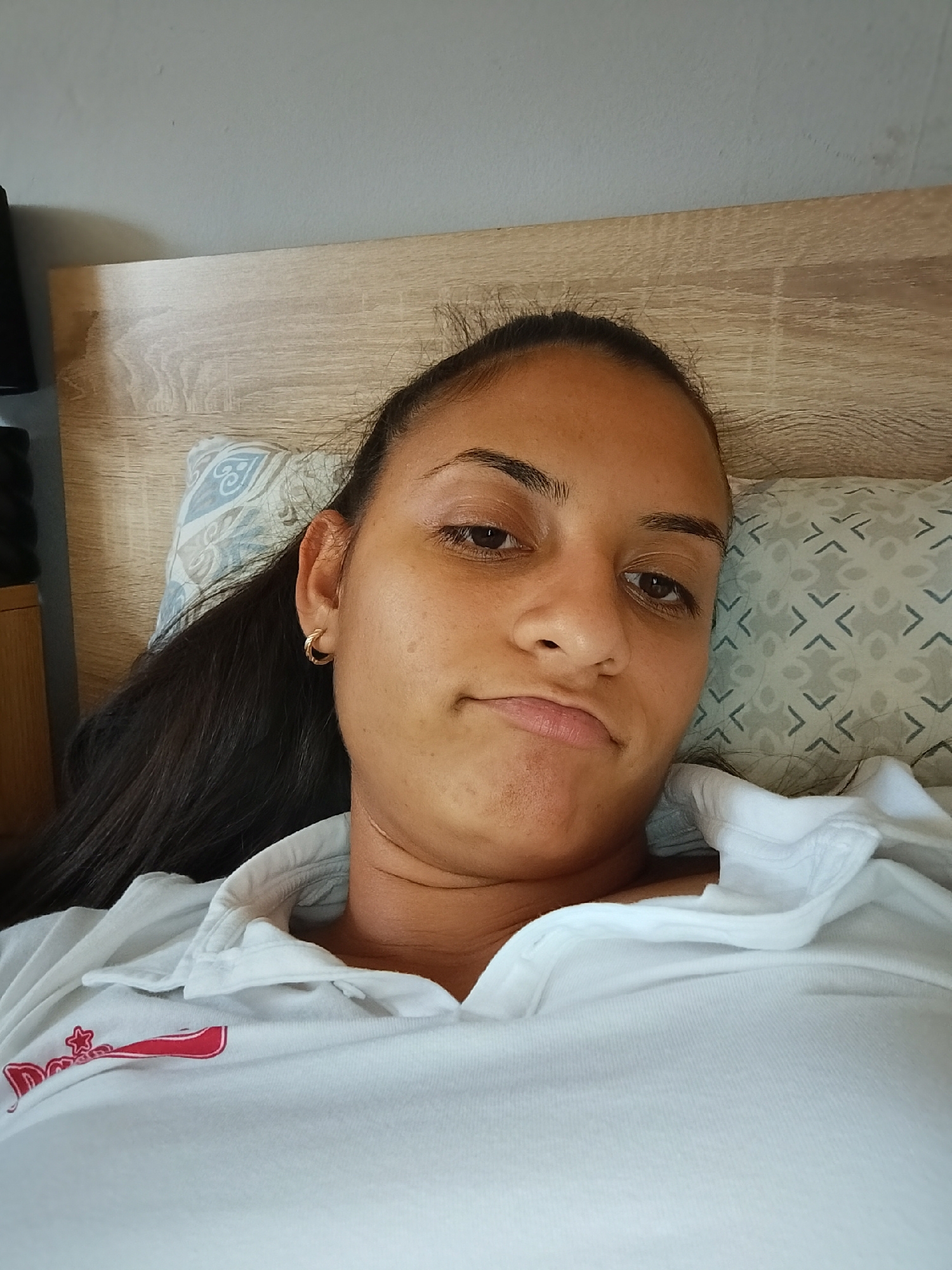 A young woman with dark hair lies on a bed. She is wearing a white collared shirt. The background consists of a headboard and a wall. The woman is looking at the camera with a neutral expression. There is a pillow with a geometric pattern behind her head. The image appears to have been taken indoors, in a bedroom. There is no indication of the specific location in the city of Yambol, Bulgaria.