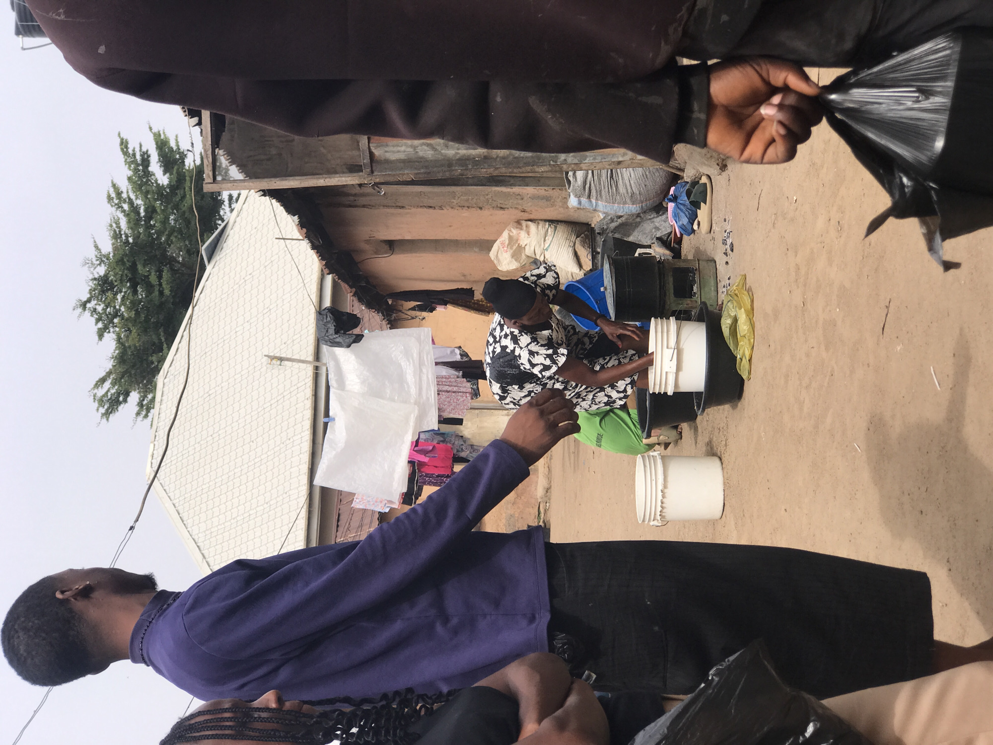 This image captures an outdoor scene within what appears to be a residential compound or yard in Jos, Nigeria, under an overcast sky during the daytime.
In the mid-ground, a person, likely female, is seated on the dirt ground, engaged in an activity involving several white, black, and blue plastic buckets. She is wearing a black and white patterned top, lime green trousers, and a black head covering. The activity suggests washing or sorting, possibly laundry, as white plastic sheets or clothes are seen hanging on a line in the background to the left.
In the immediate foreground, from an overhead perspective, a person wearing a dark brown garment is visible on the right, holding a black plastic bag in their right hand. To the left foreground, another individual is seen from behind, wearing a purple long-sleeved shirt and dark trousers, looking towards the activity in the mid-ground. Part of another person's legs, wearing light-colored trousers, is visible in the lower right corner.
The setting features unpaved dirt ground. In the background, various household items are visible, including sacks (one white sack has faint black text, possibly "RAW"), blue fabric, and footwear (sandals). Behind these items are simple, single-story structures with walls that appear to be made of mud or unpainted concrete, topped with corrugated metal roofing. Further back, to the left, a house with a white, patterned roof is visible, with green trees towering over it against the hazy sky. A power line or cable runs across the upper left portion of the frame. The scene depicts a slice of daily life, with individuals engaged in routine tasks in a modest community setting.