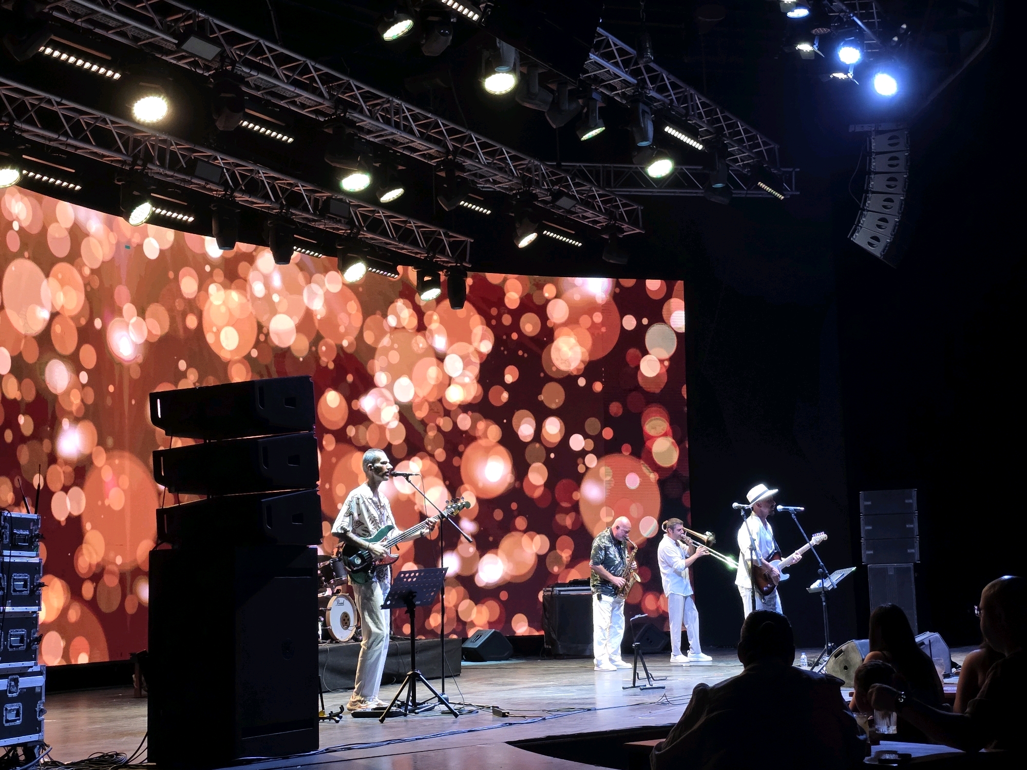 The image depicts a live musical performance on a stage with a vibrant, bokeh-effect backdrop. A band of four musicians is actively playing. On the left, a man in a patterned shirt and light pants plays a green electric guitar and sings into a microphone. Behind him, a drum kit is visible. To his right, a bald man in a patterned shirt and white pants plays a saxophone, and next to him, a man in a white shirt and pants plays a trombone. On the far right, a man in a white shirt, white pants, and a white hat plays a guitar and sings into a microphone.
Several large black speakers are stacked on the stage, and smaller equipment cases are visible to the left. Stage lighting rigs with numerous lights are suspended above the performers. The stage floor is visible, with cables running across it. In the foreground, the backs of several audience members are visible, seated at tables, with drinks on them.
The backdrop features a large LED screen displaying a pattern of out-of-focus orange and pink circles of varying sizes. The overall ambiance suggests an indoor concert venue. This scene is from the Halikarnas Club, a well-known music venue in Bodrum, Turkey, known for hosting live performances.