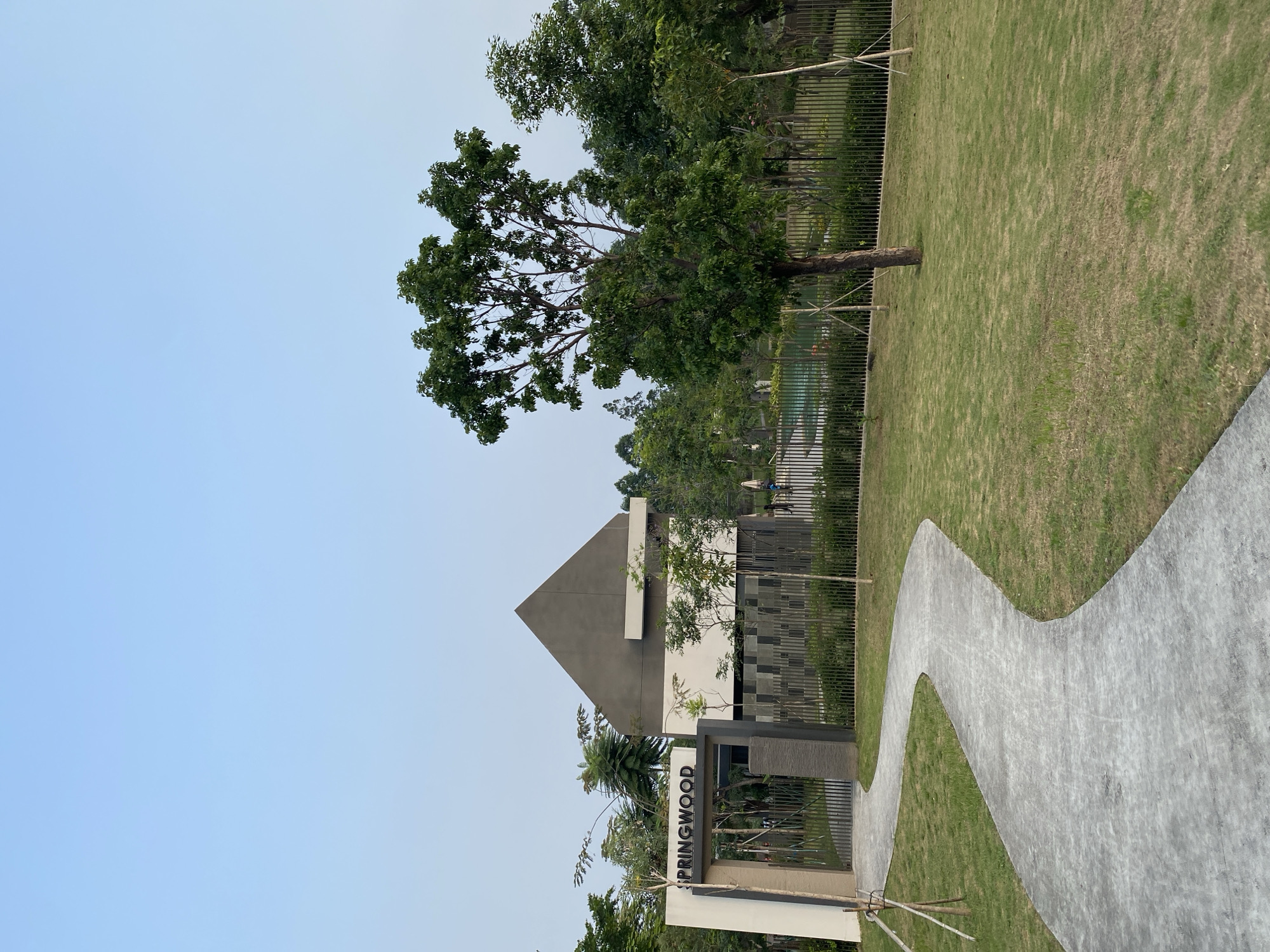 The image captures the entrance to Springwood in North Jakarta, Indonesia. A winding gray pathway leads towards a modern architectural structure with a peaked gray roof and white walls. To the left of the pathway is an entrance archway with the name "SPRINGWOOD" prominently displayed in black lettering. A black metal fence runs along the perimeter of the property. Beyond the fence, lush green trees and some landscaping are visible, along with a glimpse of a swimming pool. The sky above is a clear, pale blue, suggesting it's either early morning or late afternoon. A few young trees with stakes for support are planted in the grassy areas. The overall scene depicts a well-maintained residential or commercial development entrance.