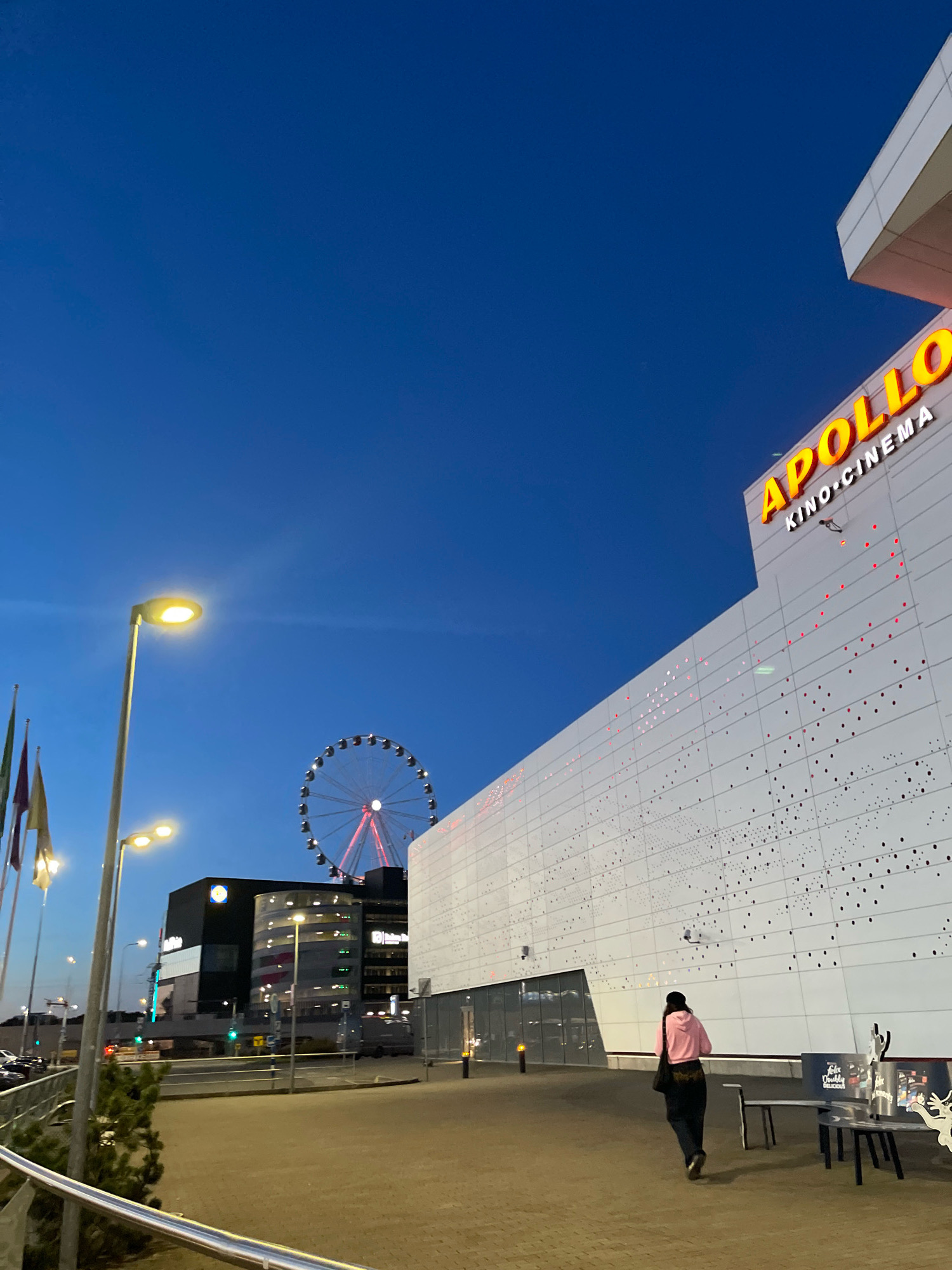 The image shows an outdoor scene at dusk in Tallinn, Estonia. In the foreground, a paved walkway leads towards a modern white building with the illuminated "APOLLO KINO CINEMA" sign. The building's facade is decorated with a pattern of red and white dots. A person wearing a pink hooded jacket and dark pants is walking away from the camera towards the building. Several benches are visible in front of the building. To the left, a street light illuminates the walkway. In the background, a large Ferris wheel with red lights is visible, along with other buildings, including a dark building with illuminated windows. Flags are also seen on flagpoles to the left. The sky is a deep blue, indicating twilight.