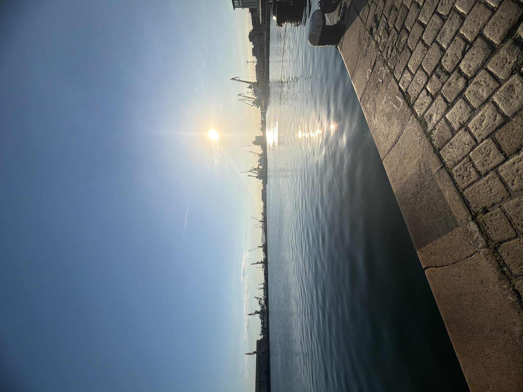 The image shows a waterfront scene, possibly a harbor or port. In the foreground, there's a cobblestone quay or walkway. The water is calm, reflecting sunlight, and in the distance, there are silhouettes of cranes and industrial structures against a bright sky. The sun is visible, creating glare and affecting the overall lighting of the scene. The perspective is taken from a low angle, near the edge of the water. The city or location is unidentified, but it appears to be an industrial or port area. There are no people visible in the shot.