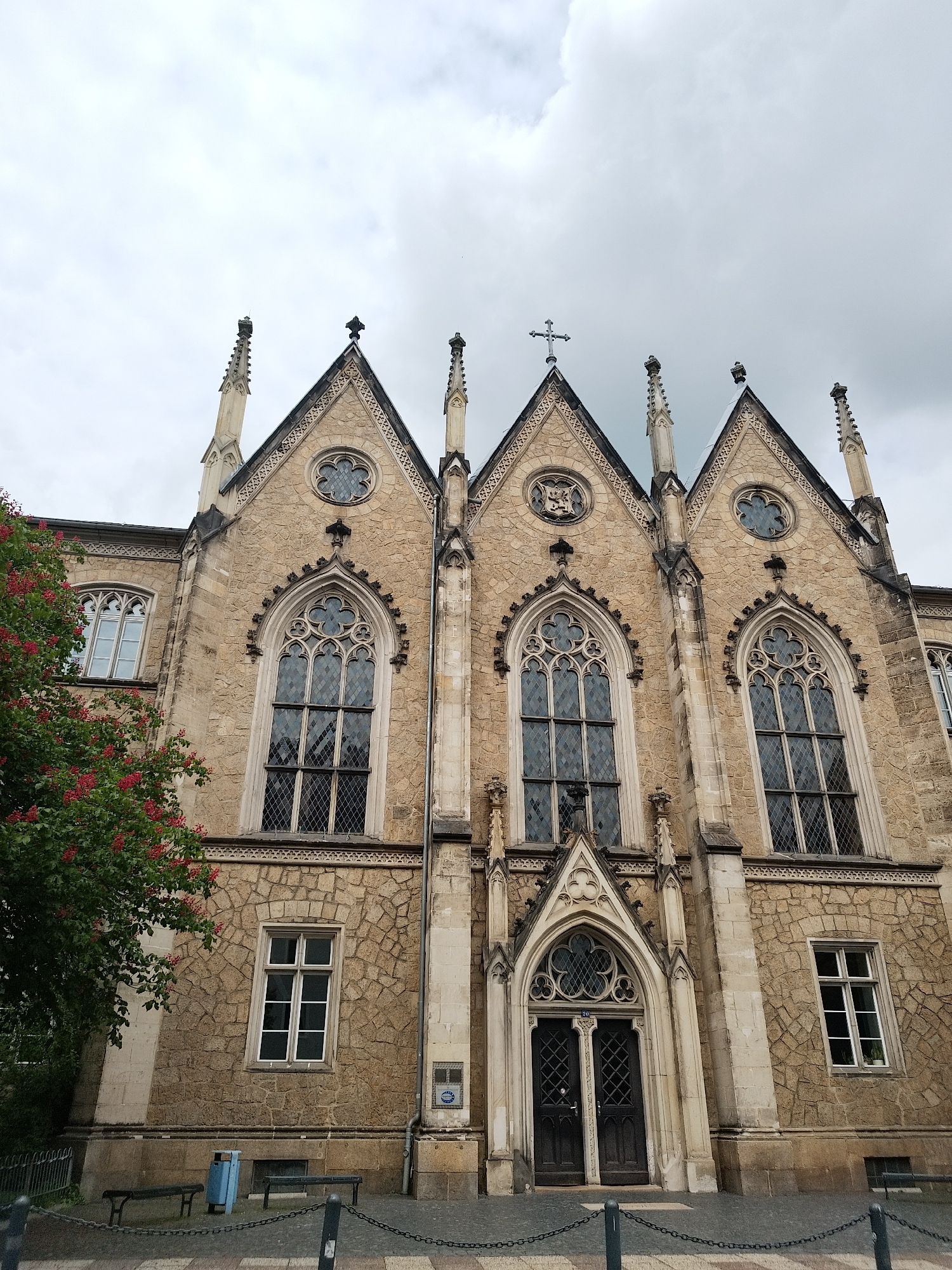 The image shows a three-peaked facade of a stone building in Unknown, Unknown, featuring gothic architectural elements. The building has three tall arched windows, a central doorway, and smaller windows to the sides. Each peak is adorned with decorative spires and a cross atop the central peak. The sky is overcast. In the foreground, there is a chain barrier, benches, and a tree with red flowers to the left.