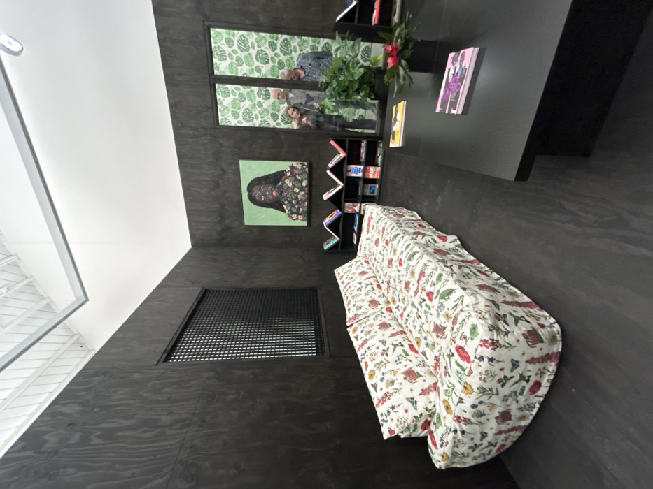 A cozy room with black walls and a floral-patterned sofa. A painting of a woman with flowers adorns the wall above a small bookshelf filled with colorful books. A table with books and a plant sits near the window with a playful view. A window with a grid-like screen and a second window with a leaf pattern can also be seen in the room.