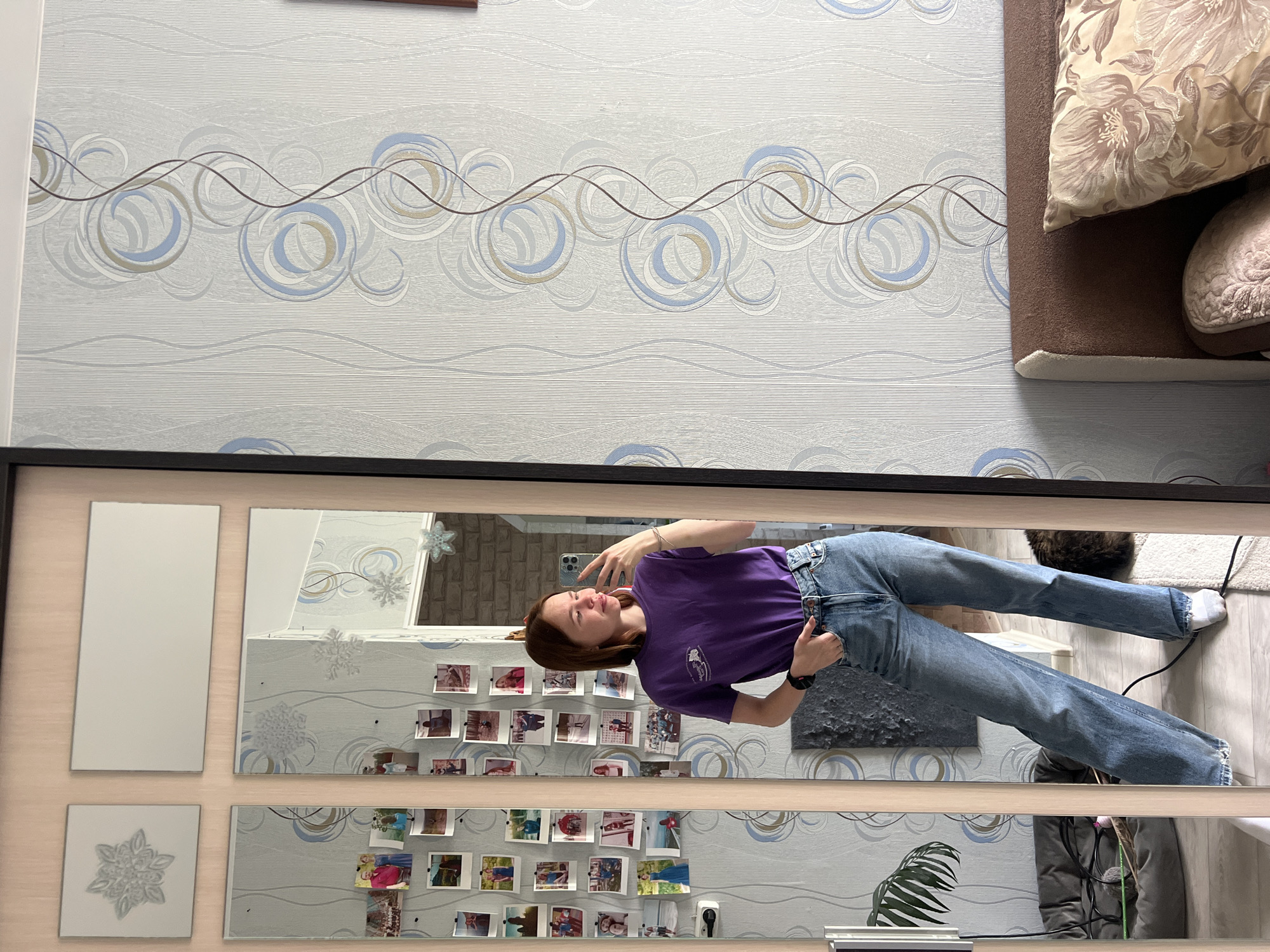 A woman is reflected in a mirror, taking a selfie in an interior space. The wall behind her has light gray wallpaper with a wave and circle pattern in blue, white, and brown. Adjacent to the wallpaper, a sofa with decorative pillows is visible. The mirror is divided into sections. Photographs are attached to the wall beneath the wallpaper in the mirror's reflection. The woman is wearing a purple t-shirt and blue jeans. She is holding a smartphone in her right hand, appearing to take a photograph. In the reflection, an electrical cord runs along the wall, and the corner of a cat bed with a cat is visible. A potted plant is also visible. The location is Chaikovsky, Russia.