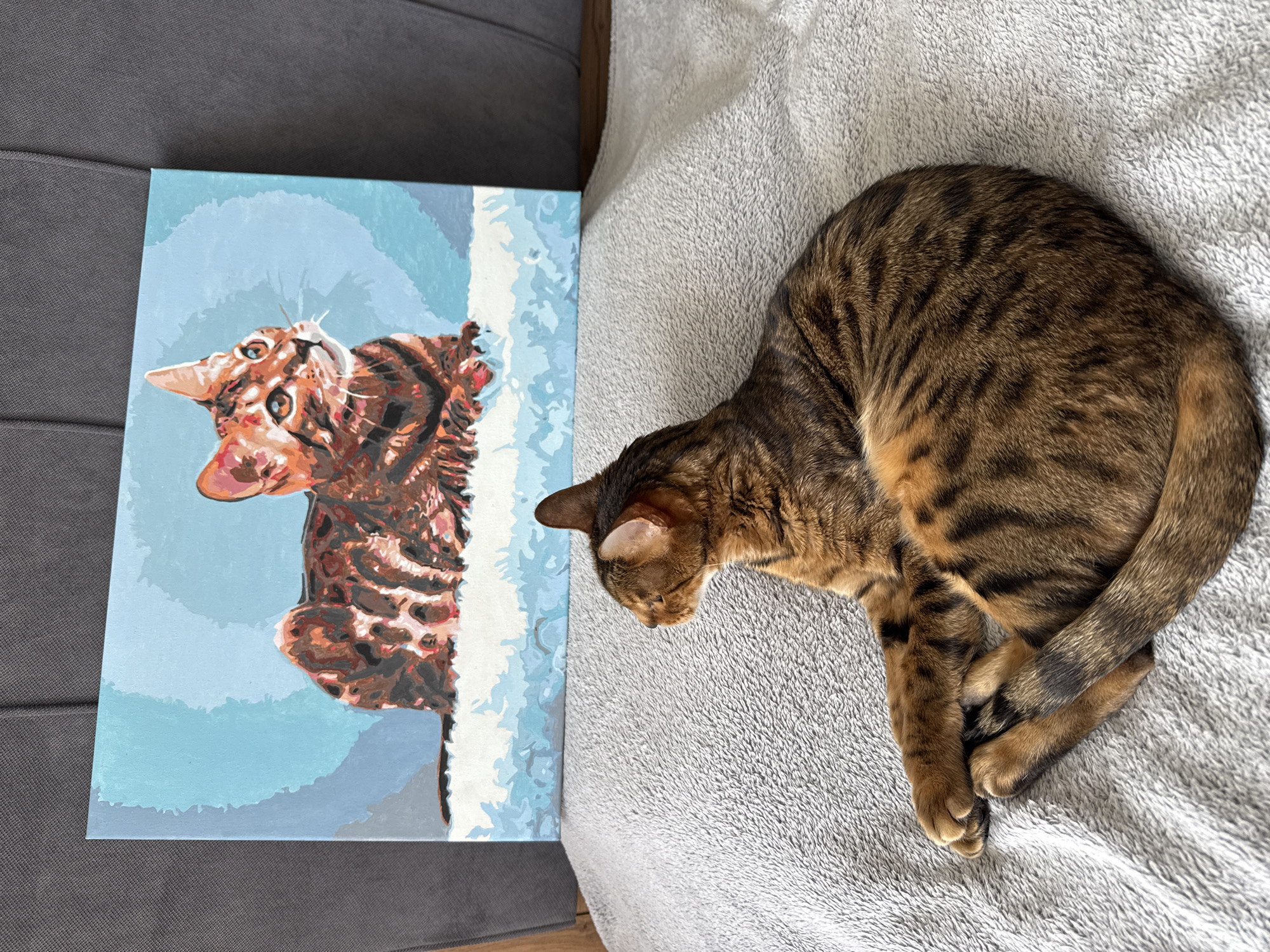 A brown tabby cat is pictured resting on a light grey, textured blanket, positioned next to a canvas painting of a cat. The real cat is curled up on its right side, its back towards the viewer, with its head turned to face the painting. Its fur displays classic tabby patterns with dark brown stripes and spots against a lighter brown and orange base.
The painting, situated to the left of the real cat, features a stylized portrait of a cat's head and upper body. The painted cat has similar brown and black tabby markings, with bright orange and lighter brown patches on its face, and is depicted looking upwards and to the left. The background of the painting consists of various shades of blue, with abstract white shapes on the right side. The painting rests on a darker grey, ribbed surface, likely a sofa or cushion. The scene is brightly lit by natural light, suggesting daytime. No discernible text is visible in the image.