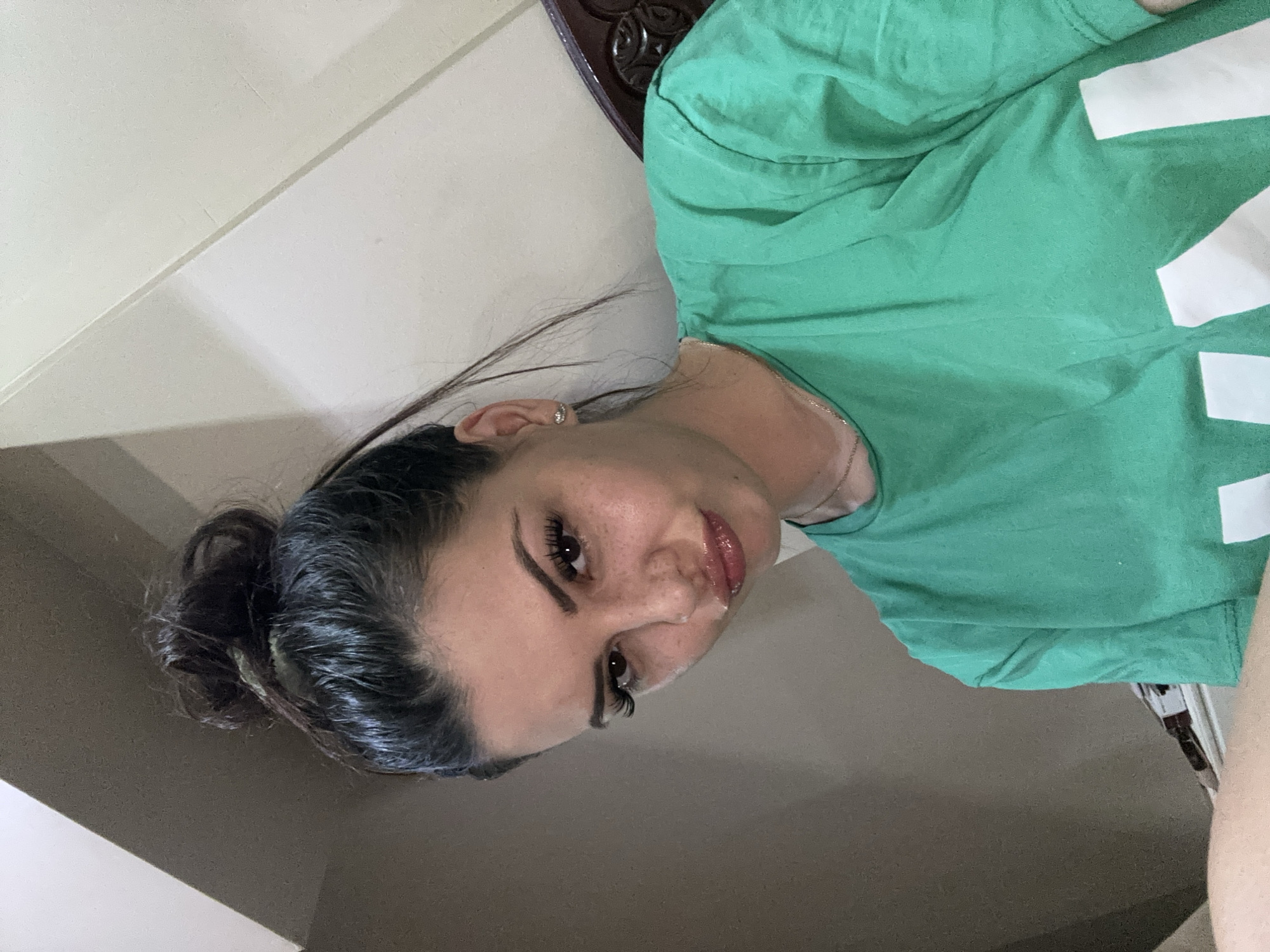 A young woman with dark hair pulled into a messy bun and light-colored freckles is looking directly at the camera with a pouty expression. She is wearing a bright green t-shirt. The background appears to be indoors with a wall that has a corner. The lighting suggests it is daytime. The specific location in Sliven, Bulgaria cannot be determined from the image.