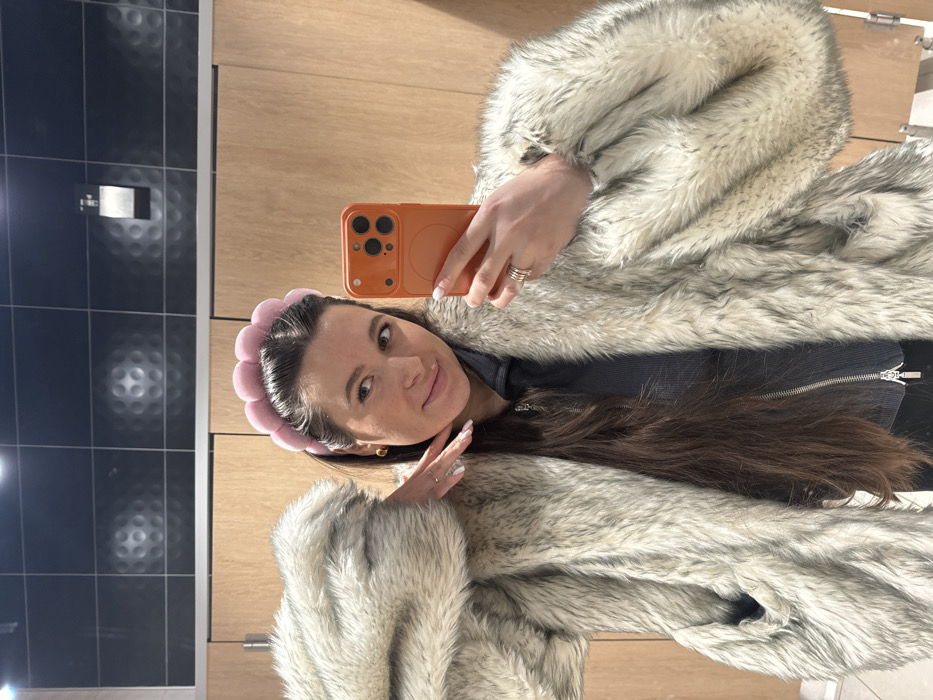 The image depicts an adult female capturing a mirror selfie in an interior setting. The subject, with long dark hair, is wearing a pink, padded headband and gold-colored stud earrings. She is dressed in a light-colored faux fur coat, primarily off-white with gray streaks, over a dark gray ribbed zip-up top. Her left hand is raised to her cheek, fingers splayed, displaying light-colored nail polish. Her right hand holds an orange-cased smartphone with a multi-lens camera module, pointed at her reflection. She wears several rings on her left ring finger. The background consists of a dark tiled wall with a repeating dimpled pattern in the upper section and lighter wooden or laminate paneling below. A white, wall-mounted toilet paper dispenser is visible on the dark tiled section. The setting appears to be a public restroom or changing area. There are no discernible visual cues within the image to confirm a specific geographical location such as Akseno-Butyrki, Russia.
