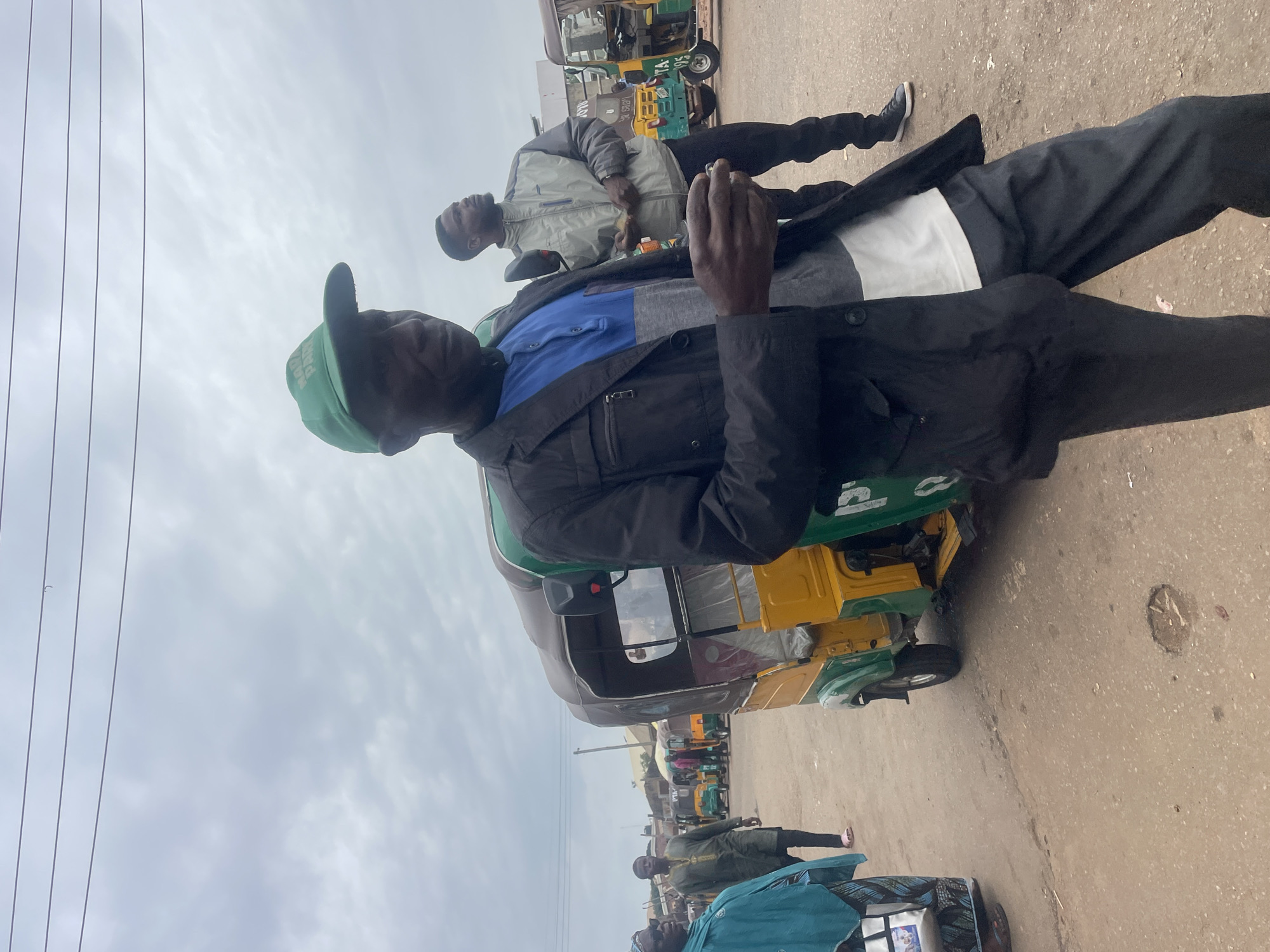 The image depicts an outdoor scene with overcast skies, likely in the city of Gura Top, Nigeria. In the foreground, a man wearing a green cap and a dark jacket over a blue and gray shirt is looking to the right, holding a small object in his right hand. Behind him, another man in a gray jacket and dark pants is also looking to the right, holding a small object. To the left of the foreground man, a yellow and green auto-rickshaw is visible, with its rear and side partially obscured by the man. Further in the background, more auto-rickshaws are parked or moving. Other individuals are present in the middle ground, including a person in a blue garment and another in a green traditional outfit. The ground is a dirt or unpaved surface, with some visible potholes and debris. The overall environment suggests a busy street or transportation hub.