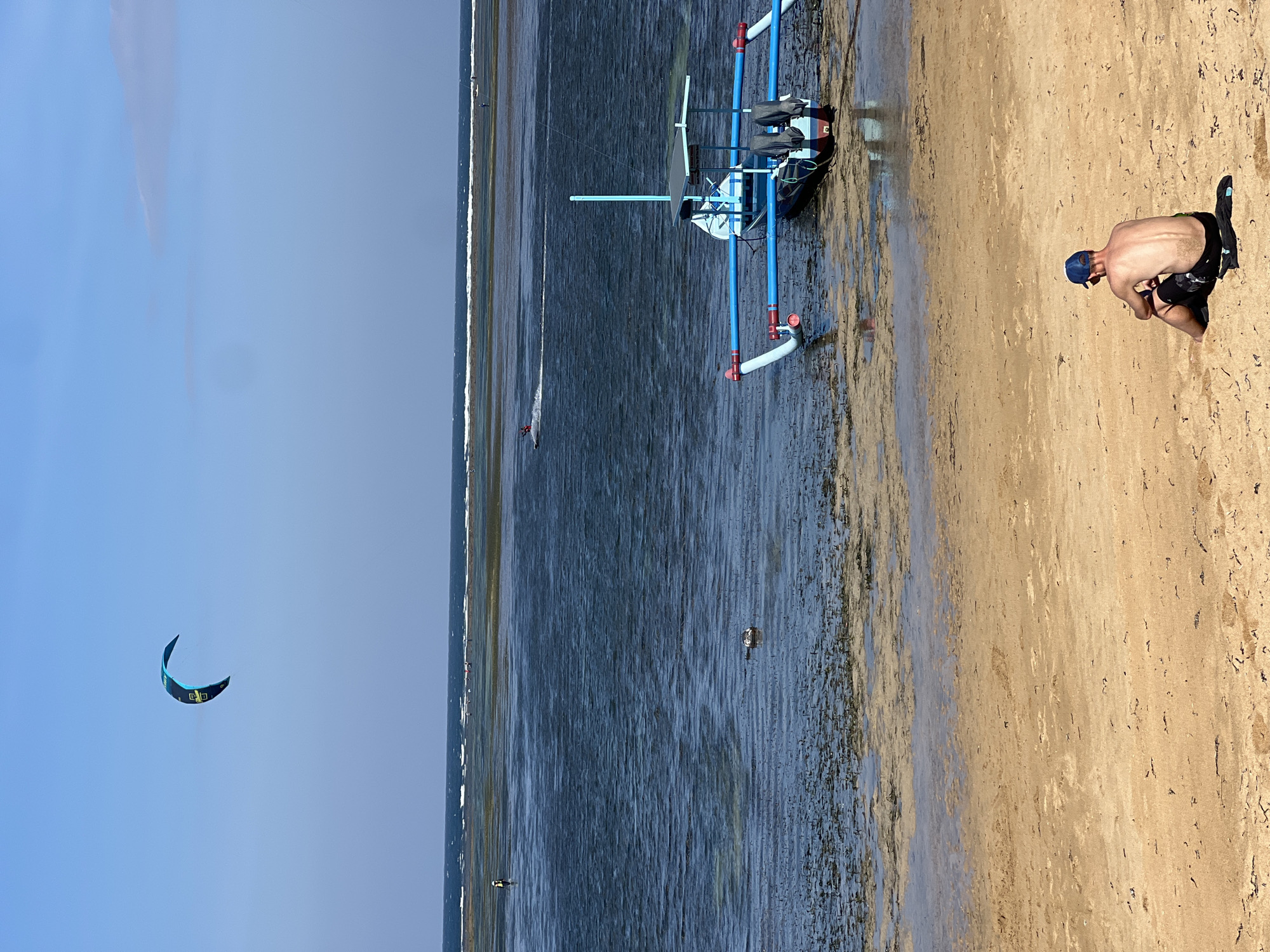 This media file depicts a scenic beach environment with elements of watersports. The scene features a sandy beach in the foreground and a body of water stretching into the distance. A man, wearing a blue cap and dark shorts, is kneeling on the sand, appearing to be engaged in some activity or rest. In the water, a person is visible kiteboarding, with a colorful kite in the sky above them, indicating an active watersport session. Further in the distance, a specialized watercraft, possibly designed for kiteboarding or a similar activity, is present, equipped with blue tubing and what appears to be a solar panel. The sky is a clear blue, suggesting fair weather and likely daytime. The overall setting appears to be a coastal area, with the tide out, exposing a wet sandbar. The image conveys a sense of outdoor recreation and leisure in a tropical or subtropical location, consistent with the provided location context of Denpasar, Indonesia. There is no visible text in the media file.