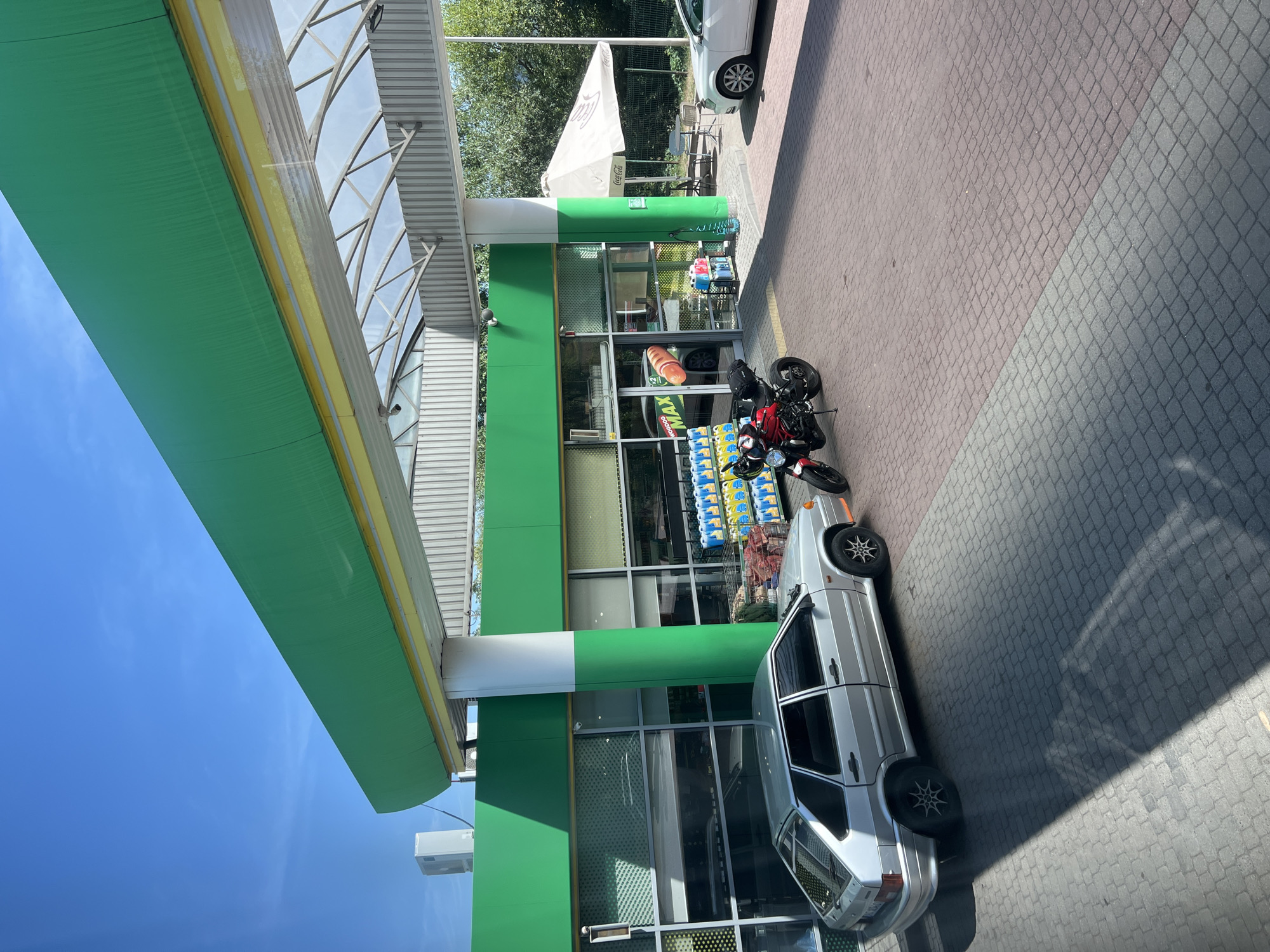 This image depicts a gas station forecourt in Chynadiiovo, Ukraine, on a clear, sunny day. A silver hatchback car is parked on the red brick paving stones, casting a long shadow. Adjacent to the car, a red motorcycle is parked. In the background, a store with large glass windows is visible, displaying rows of yellow and blue liquid containers. A green awning with a yellow trim extends over the forecourt, supported by white and green columns. Further to the right, under a large white umbrella with "Coca-Cola" branding, outdoor seating is arranged. A white sedan is parked near the umbrella. Trees line the edge of the gas station property. The sky above is a clear blue.