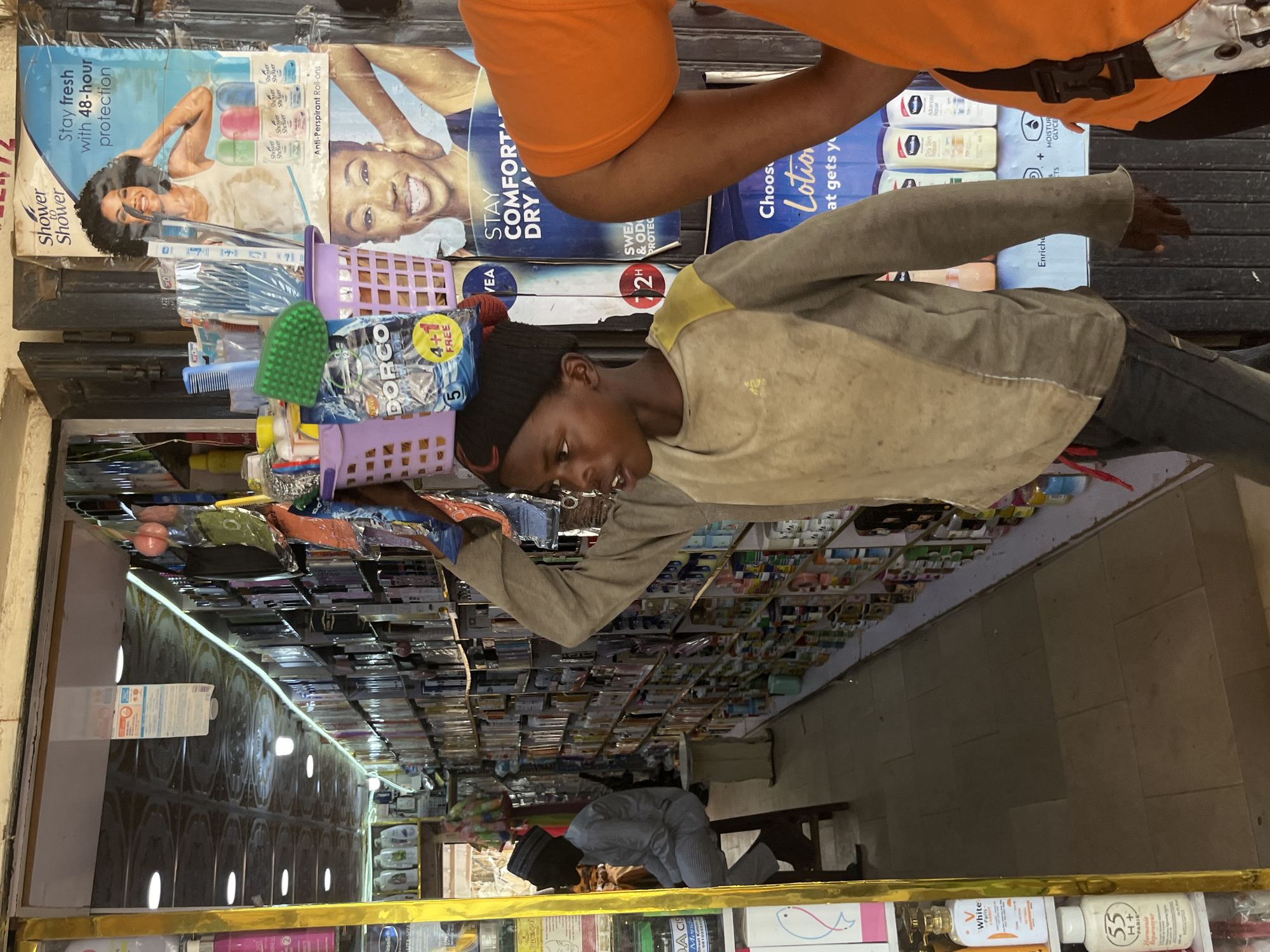 A young boy, wearing a dark beanie and a dirt-stained tan and grey long-sleeved shirt, carries a purple basket filled with various items, including a green scrub brush, on his head. He is walking through a store in Jos, Nigeria, with shelves stocked with toiletries and personal care products lining the walls. To his right, a poster advertises "Shower to Shower" anti-perspirant roll-ons and lotions. Another person, wearing an orange shirt, is partially visible in the foreground. A man in a light blue traditional outfit and a cap sits on a stool further inside the store. The store's floor is tiled, and the ceiling appears to be covered in patterned panels with fluorescent lighting.