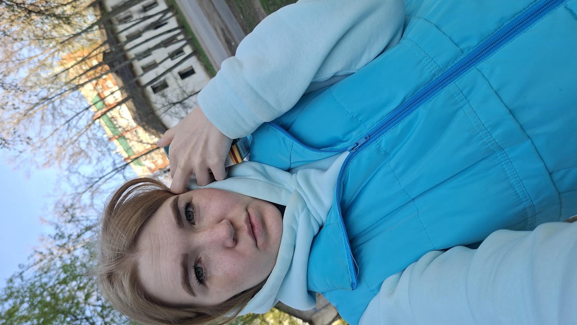 A young woman with fair skin and light brown hair takes a selfie outdoors. She is wearing a light blue hooded sweatshirt under a brighter blue quilted vest. Her right hand is near her forehead. In the background, out of focus, is a multi-story building with green and orange accents. Bare tree branches are visible in the upper left, suggesting it might be early spring. The image was taken in Chaikovsky, Russia.