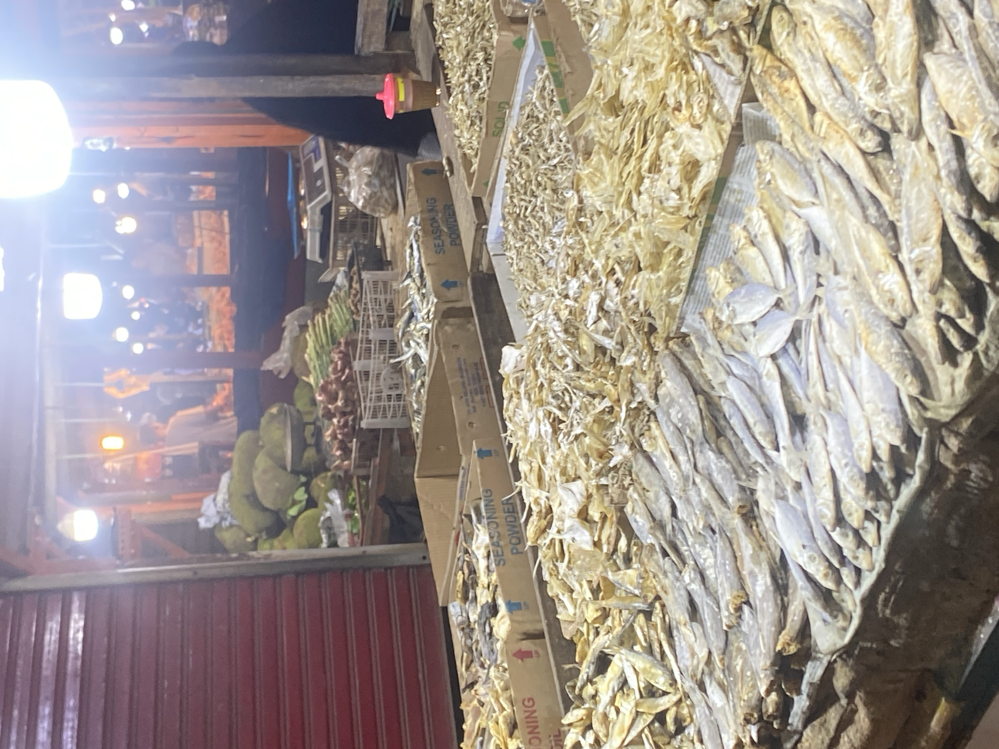 This is an outdoor market scene in Cikupa, Indonesia, featuring a large display of dried fish. The foreground is dominated by several containers of dried fish, arranged in neat rows and piles. The fish vary in size, with some appearing to be small, whole specimens, while others are larger and flatter. Behind the fish, there are cardboard boxes with text indicating "SEASONING POWDER" and "SEASONING OIL". In the background, a person wearing dark clothing is visible behind a stall. Other market elements include stacks of jackfruit, baskets of produce such as onions and green vegetables, and a weighing scale. The market appears to be lit by artificial lights, suggesting it might be evening or indoors with artificial lighting. A red corrugated metal shutter is visible on the left side of the image.