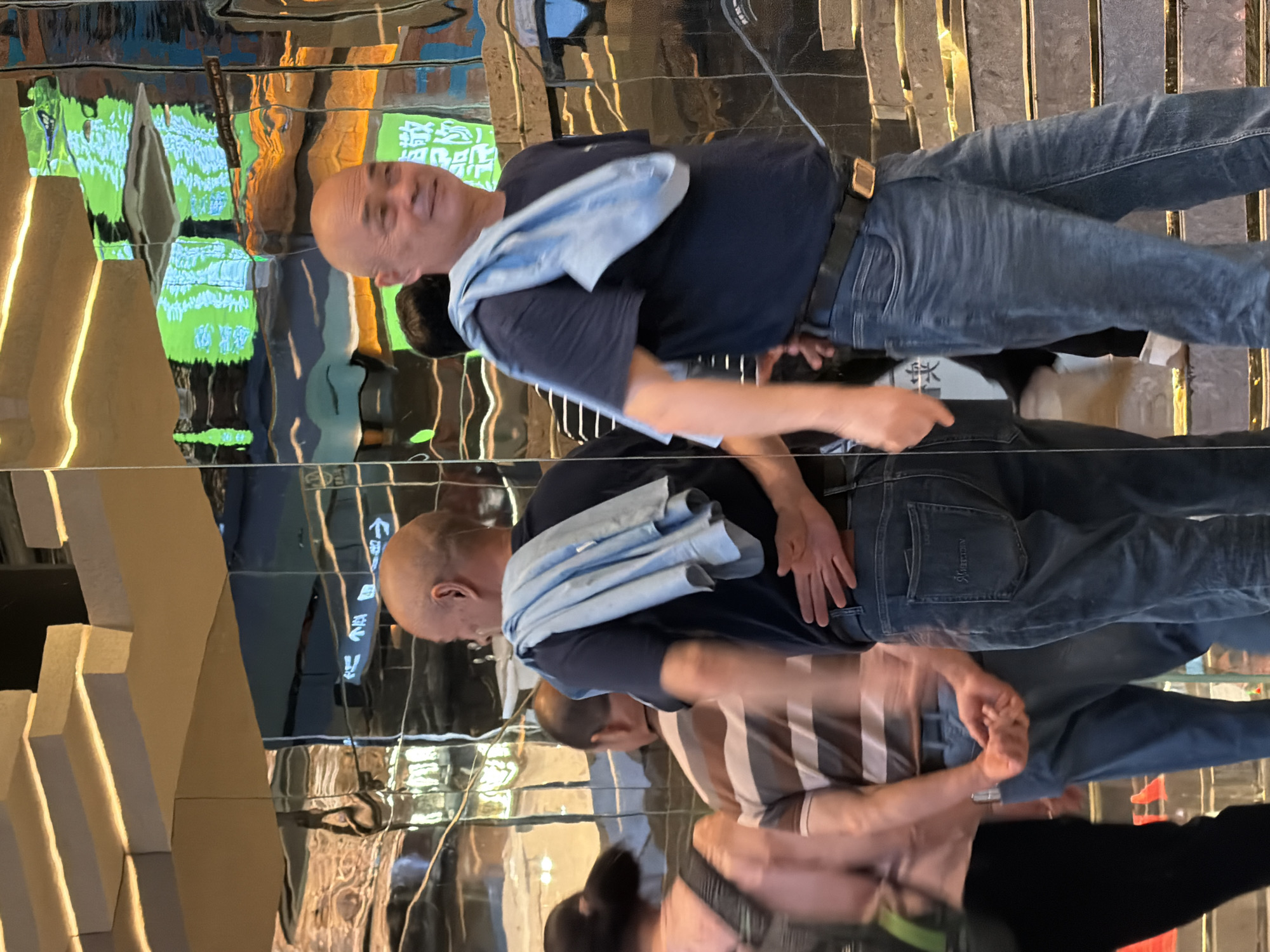 In Yuzhong District, China, two men and a woman are in a reflective environment, possibly a shopping mall. The foreground features a bald man in a dark blue t-shirt and jeans, with a light blue towel draped over his shoulders. He is looking to his right, with his right hand behind his back, and his left hand resting on his left hip. To his left, a man in a striped t-shirt and jeans is walking away from the camera, holding his hands clasped behind his back. Further to the left, a woman in a pink top and black pants with a backpack is blurred by motion as she walks away. The background consists of highly reflective, rippling surfaces that distort the reflections of the surroundings, including overhead lighting and possibly directional signs. The floor appears to be a staircase with golden and reflective accents.