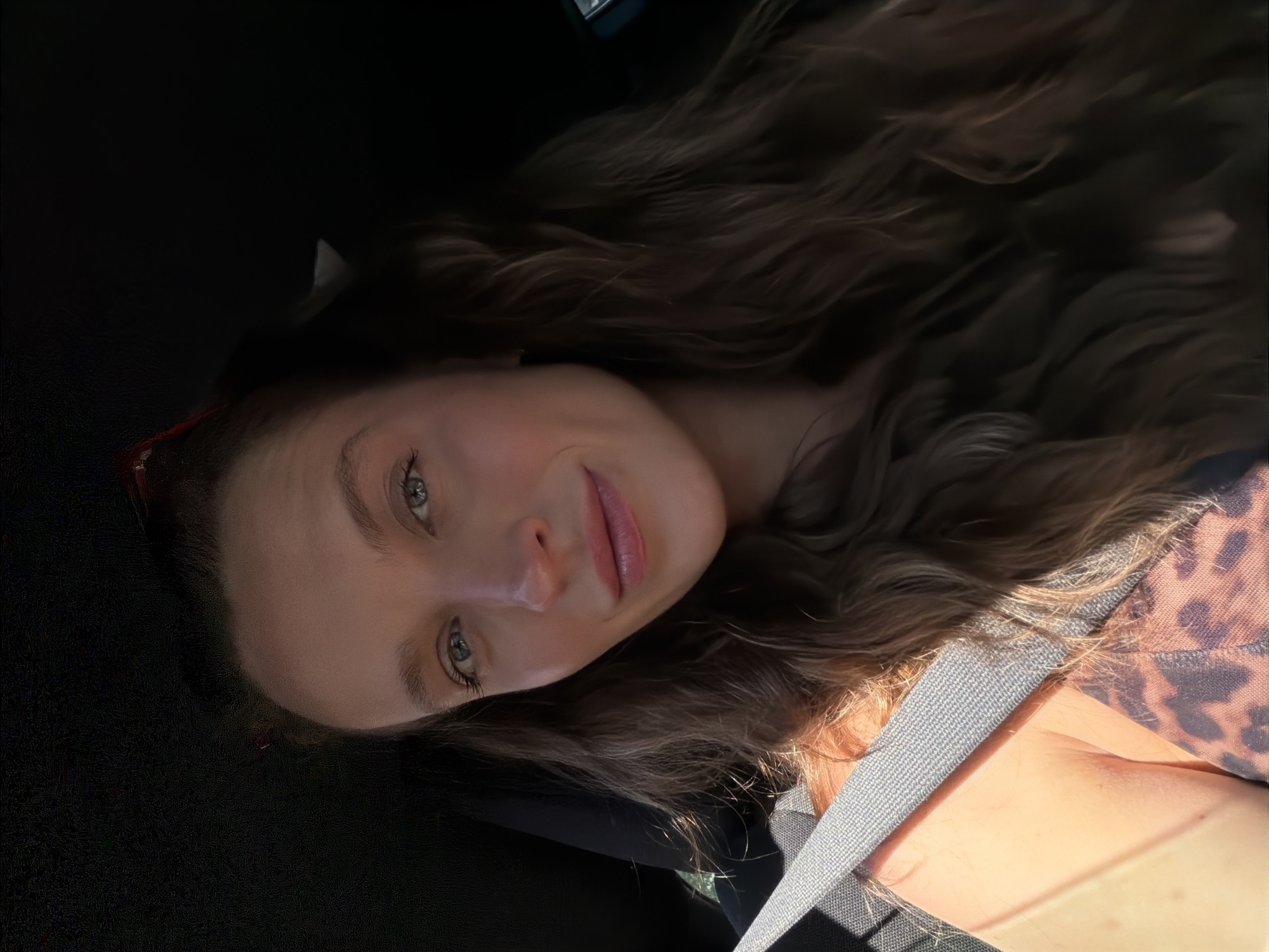 A young woman with long, wavy, brown hair and fair skin is shown in a selfie taken inside a vehicle. She is wearing a seatbelt and a garment with a leopard print pattern. The scene is partially illuminated by sunlight. The location in the city of Budva, Montenegro is not discernible from the image.